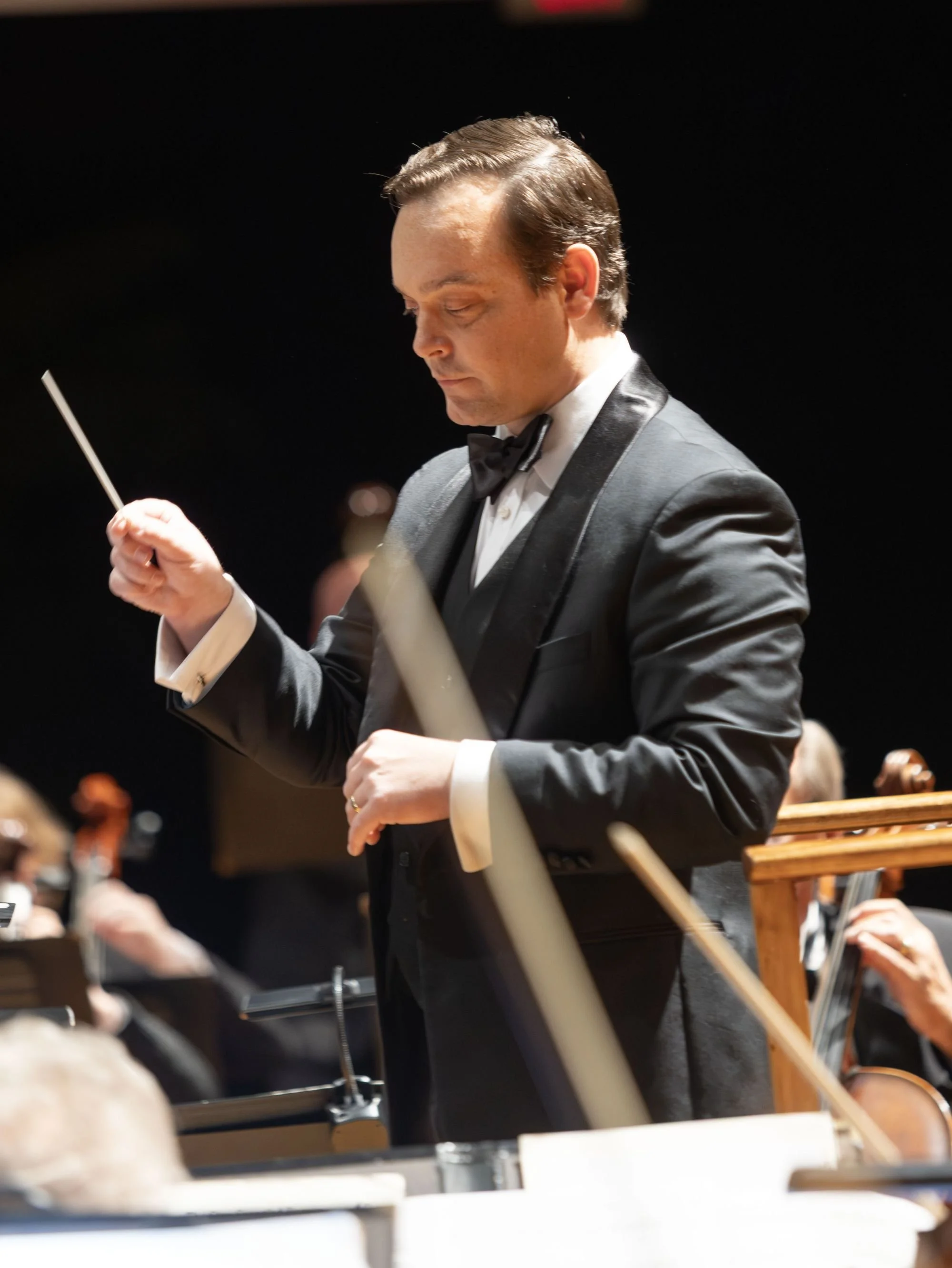 A young man in a tuxedo conducting an orchestra with a baton in his right hand.