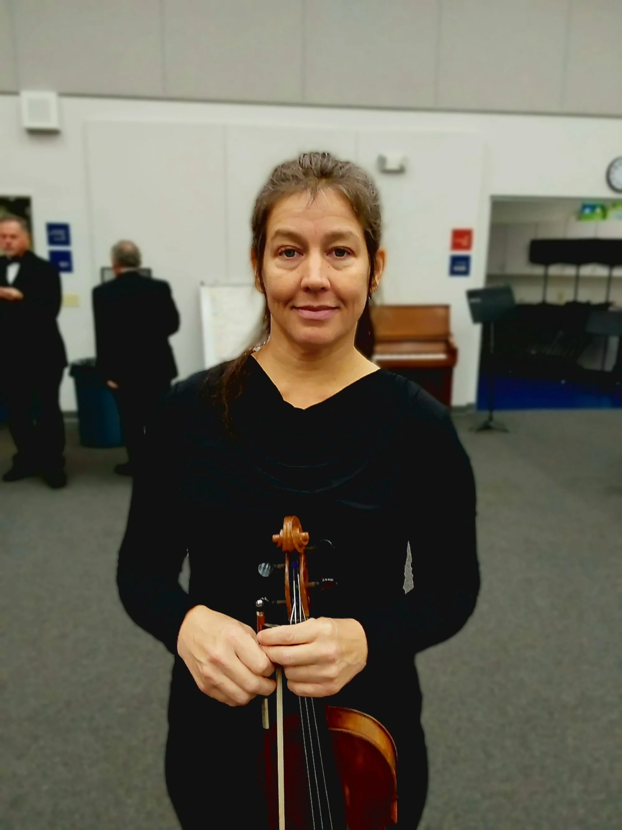 Woman in black dress holding a violin, standing in a music room with two men in tuxedos in the background.