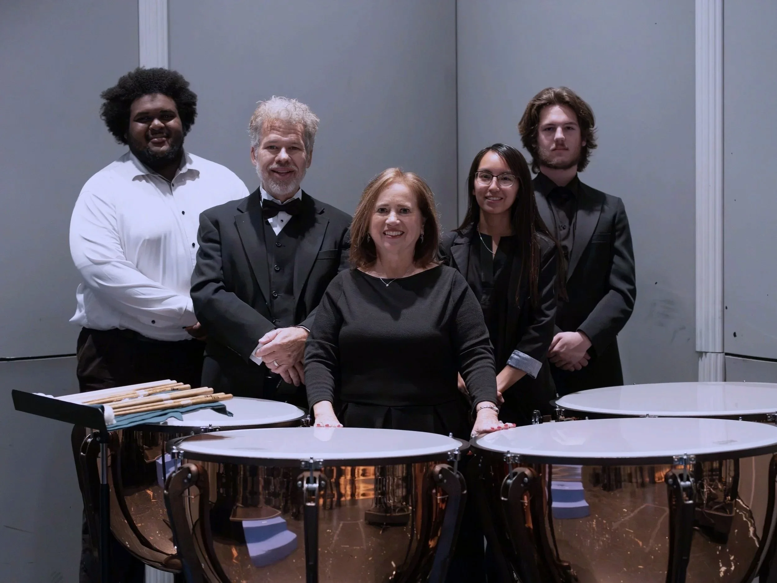 A group of five musicians in formal attire standing behind timpani drums in a concert hall.