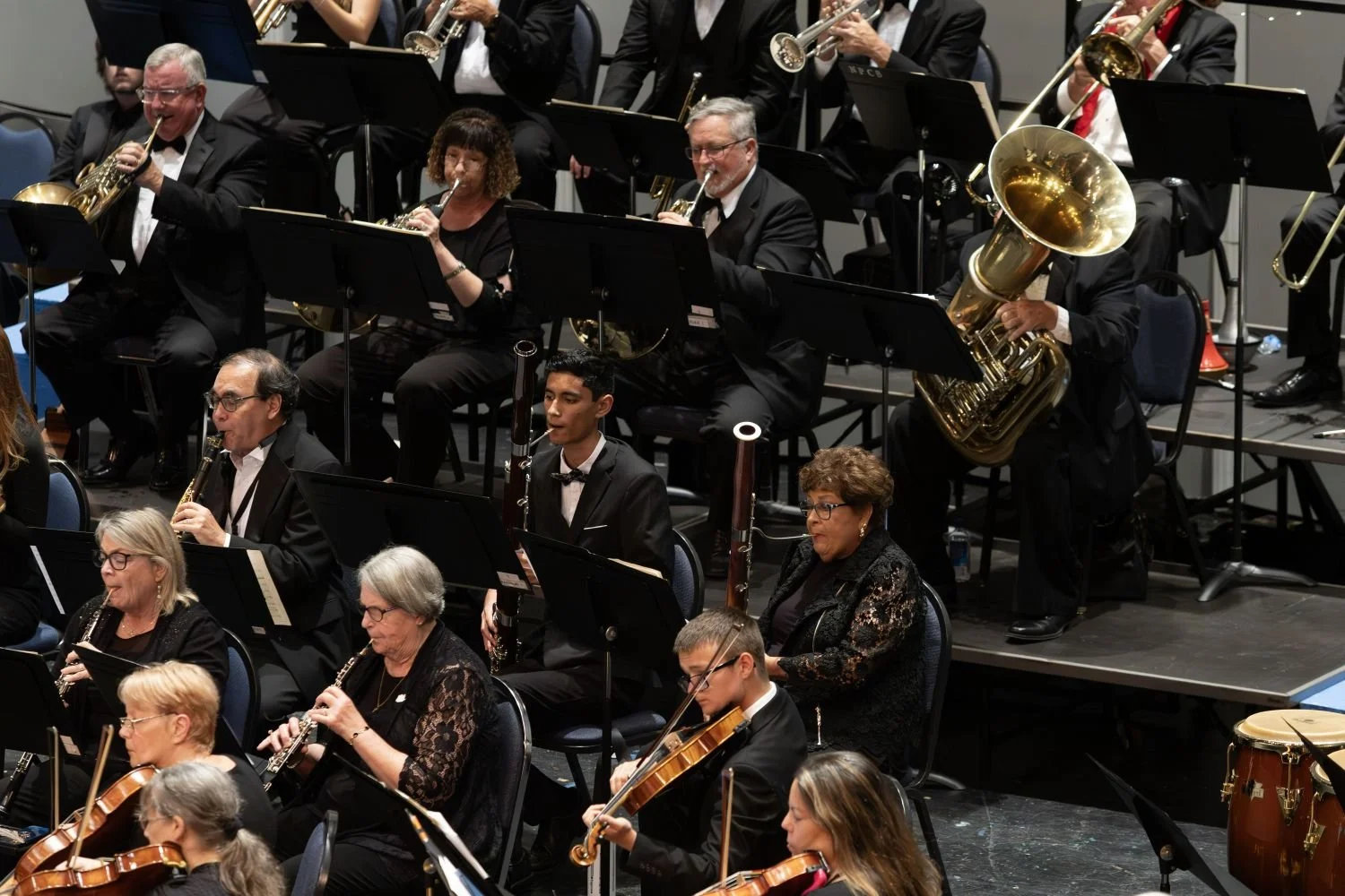 Orchestra musicians performing on stage, dressed in formal black attire, with various musical instruments including trumpets, clarinets, and violins.