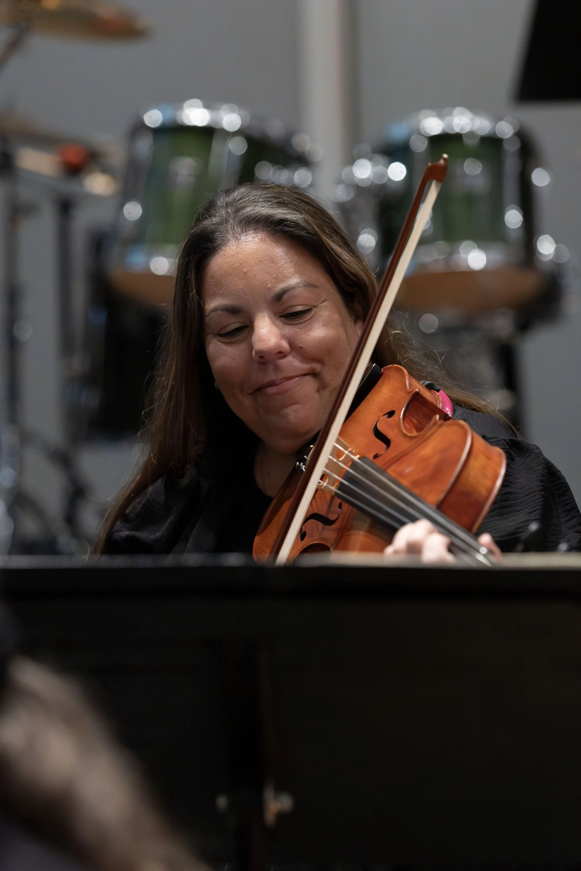 Woman playing the violin during a performance, with drums blurred in the background.