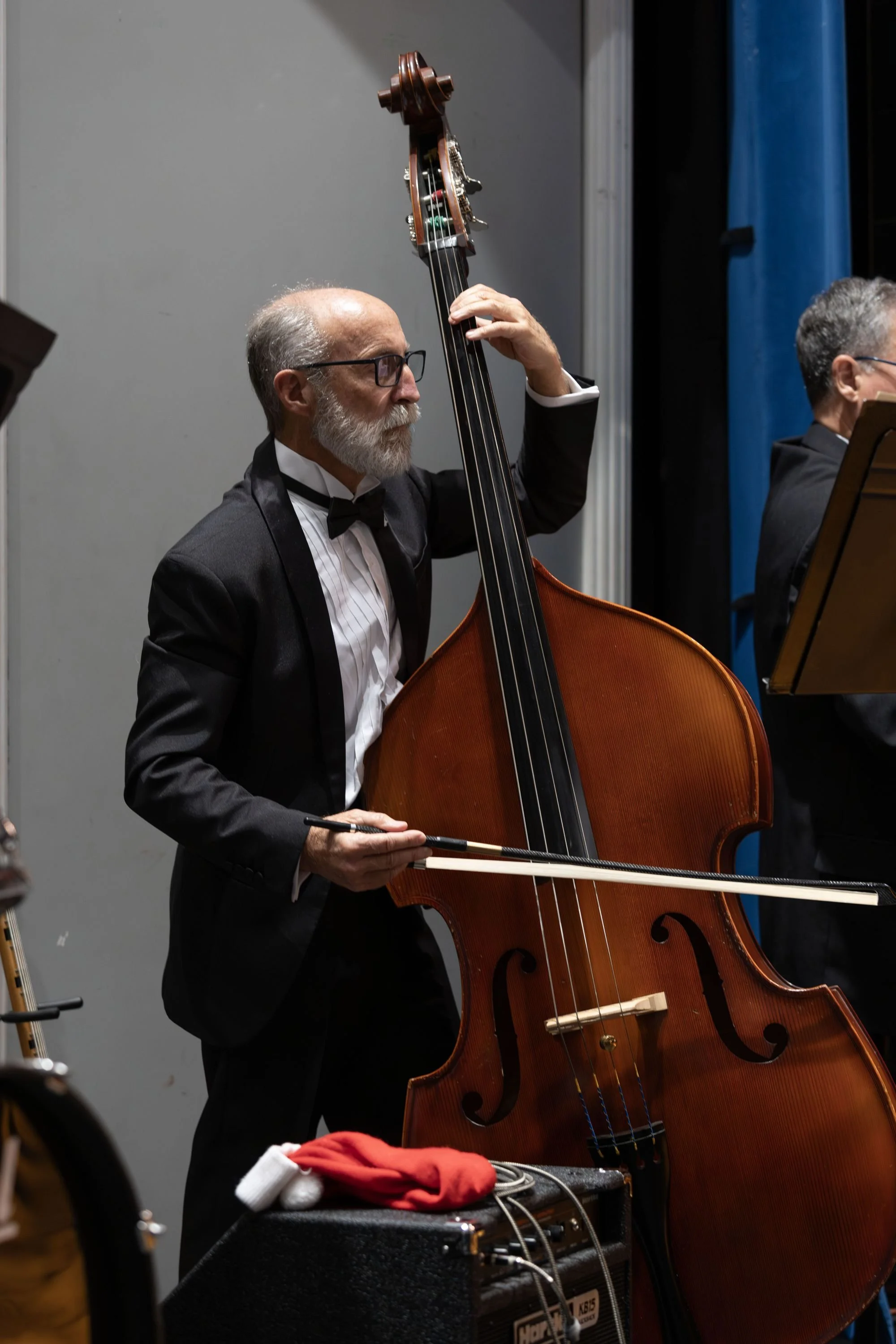 A man with glasses and a beard dressed in a tuxedo playing a double bass during a performance.