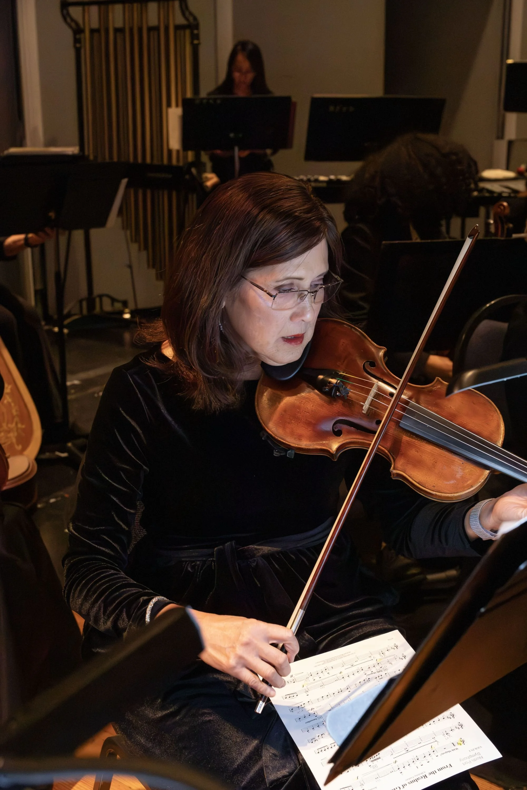 A woman with glasses playing a violin while reading sheet music in a music rehearsal room. Several other musicians are visible in the background.