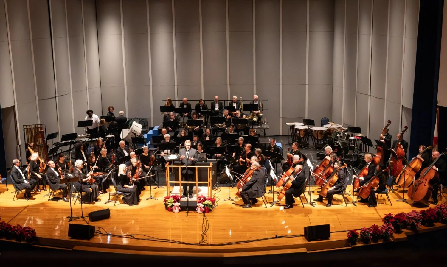 Orchestra performing on stage in concert hall with conductor and various instruments including strings, brass, percussion, and woodwinds, decorated with flowers.