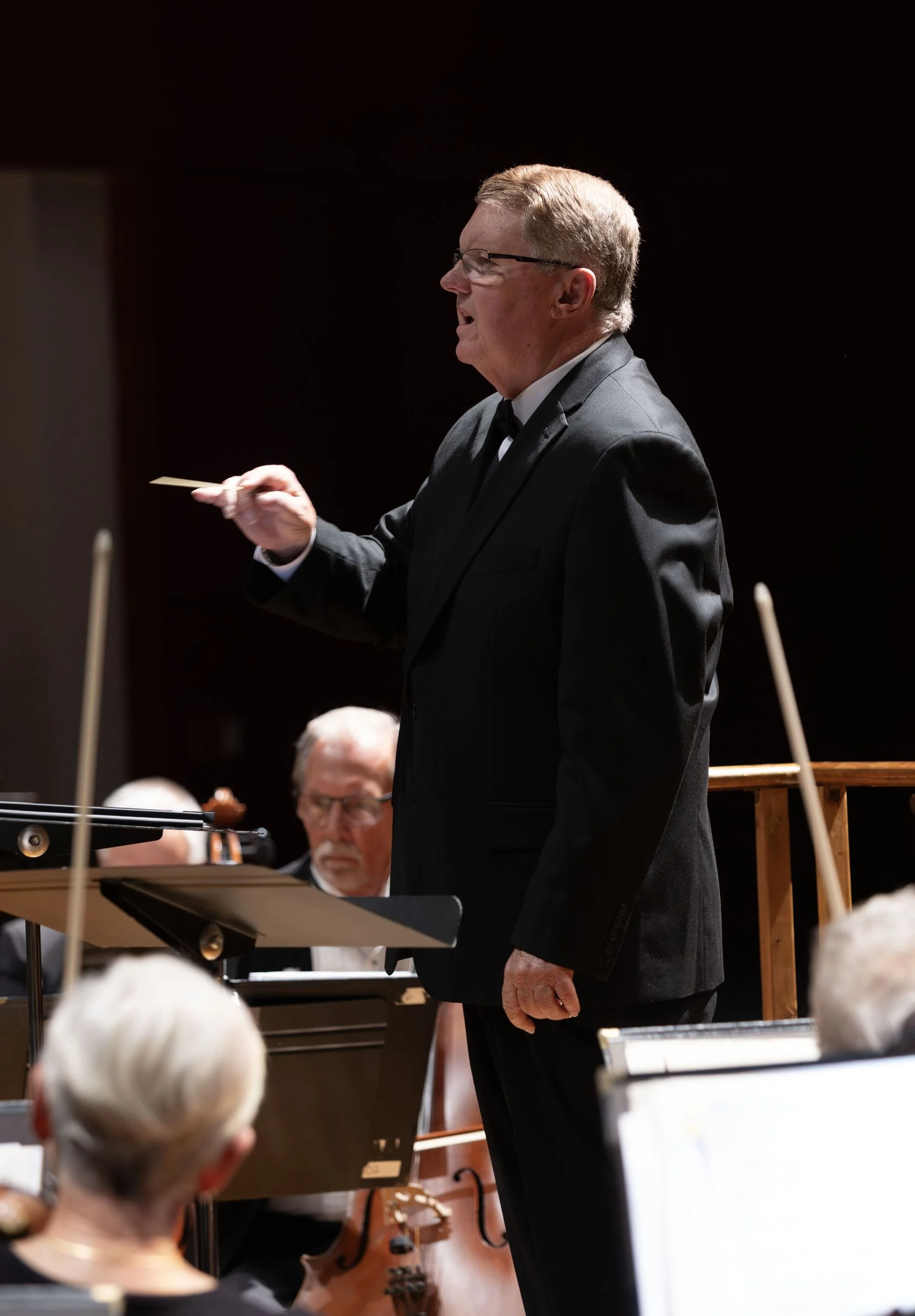 A conductor in a black tuxedo leading an orchestra during a performance.