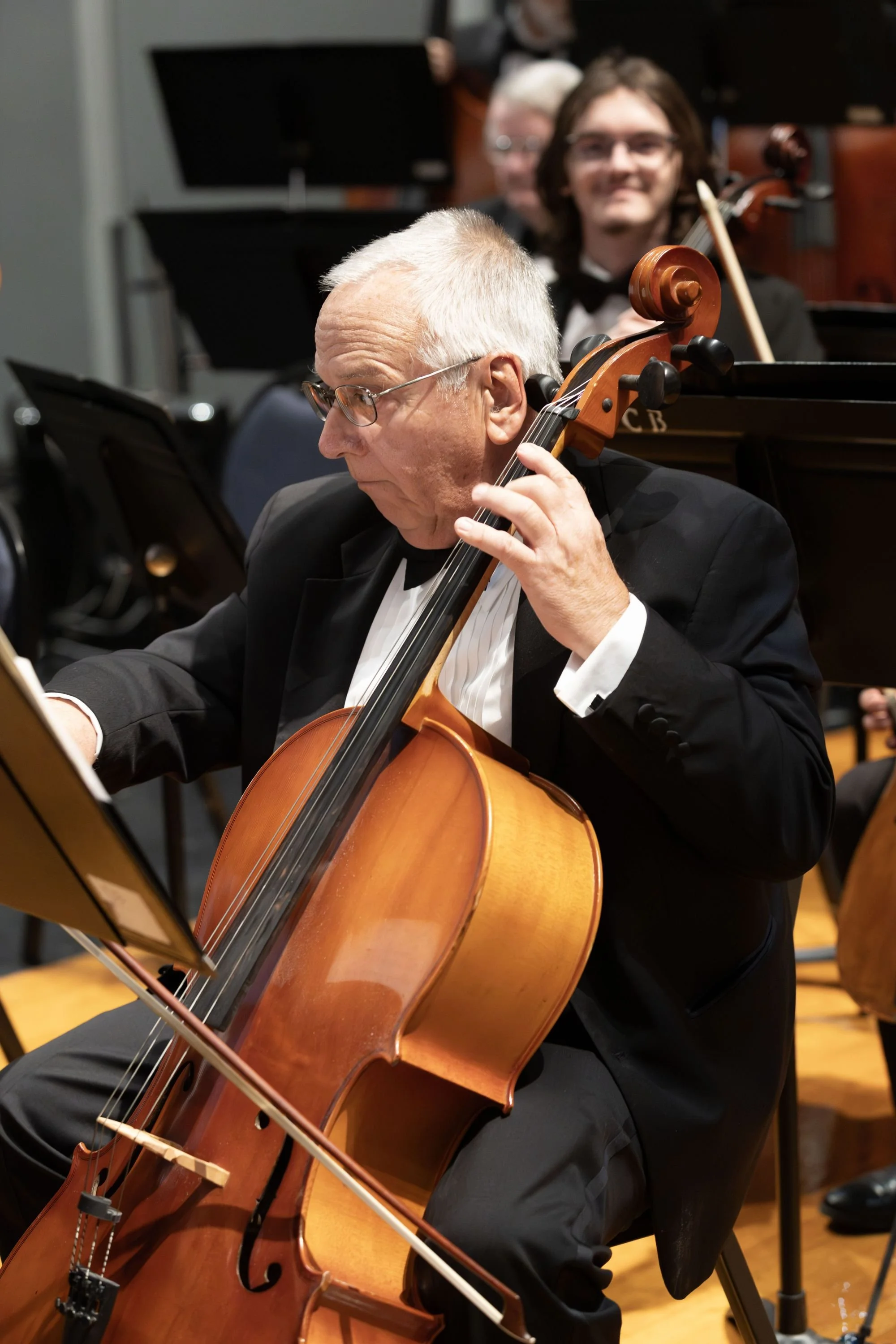 An elderly man in a tuxedo playing a cello during an orchestral performance, with other musicians in the background.
