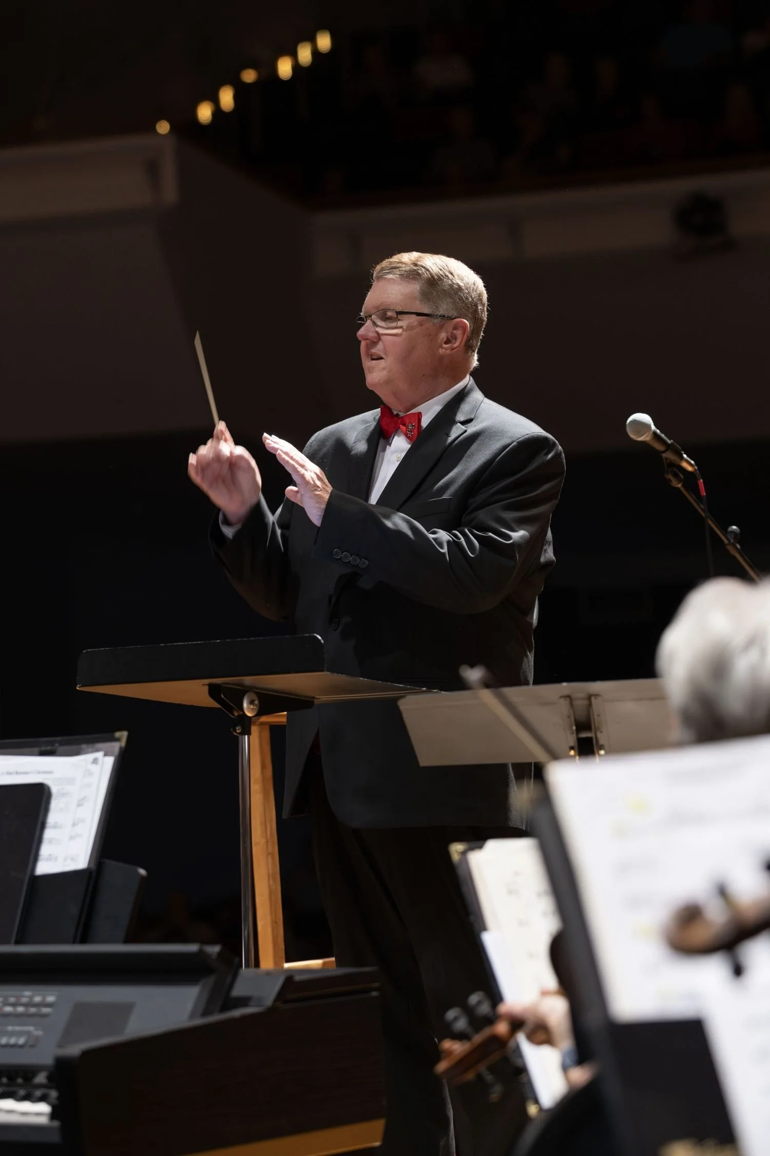 A man in a suit and red bow tie conducts an orchestra, holding a baton in his right hand, in a concert hall.