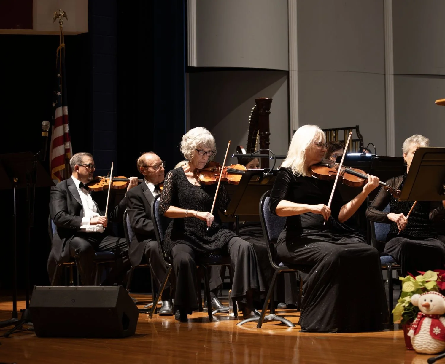 An orchestra performing on stage with string instruments, mostly violins, in formal attire, in a concert hall decorated for Christmas.