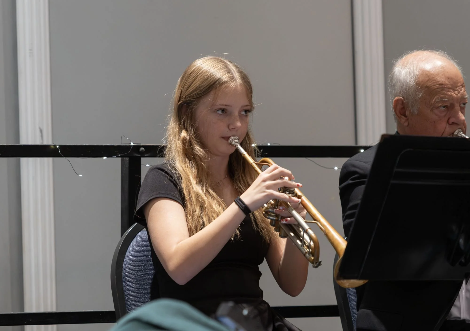A young woman with long blonde hair playing a trumpet during a musical performance, sitting next to an older man.