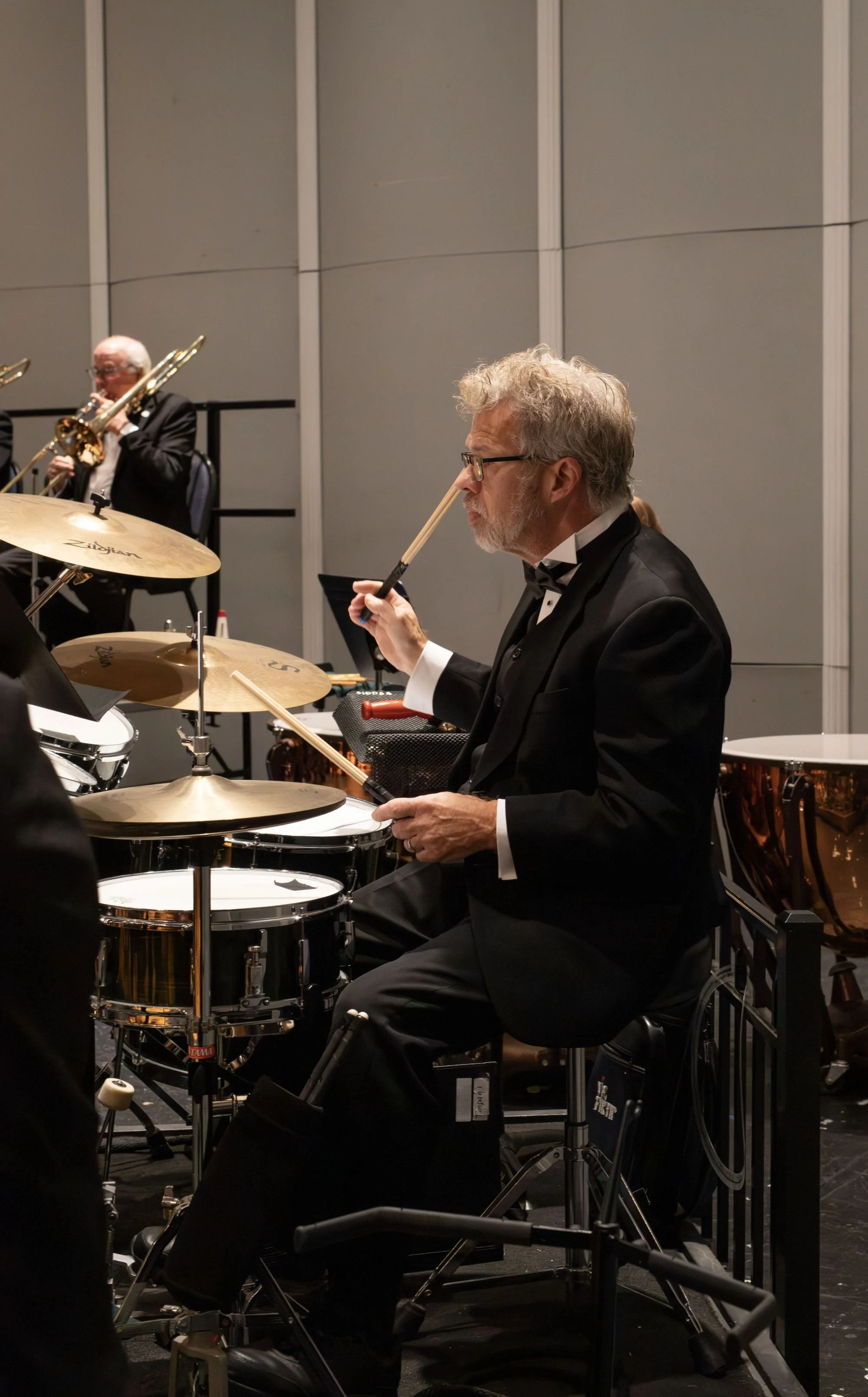 An older man with gray hair, glasses, and a beard, dressed in a black tuxedo, playing drums during a formal concert with other musicians in the background.