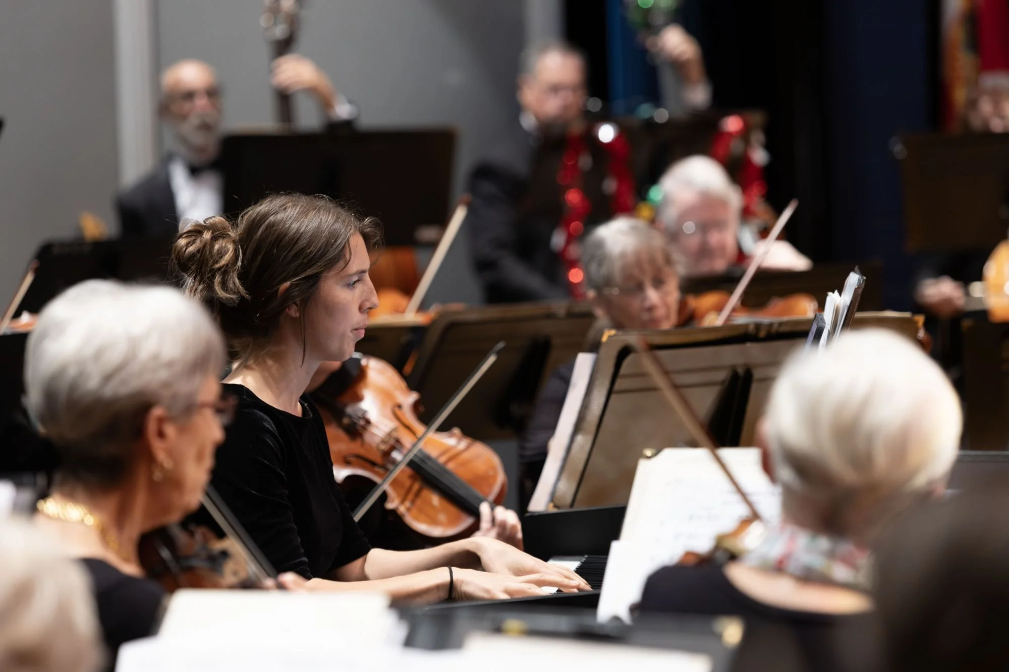 Orchestra members playing string instruments during a performance, focusing on a young woman playing the piano and others playing violins, with sheet music in front of them.