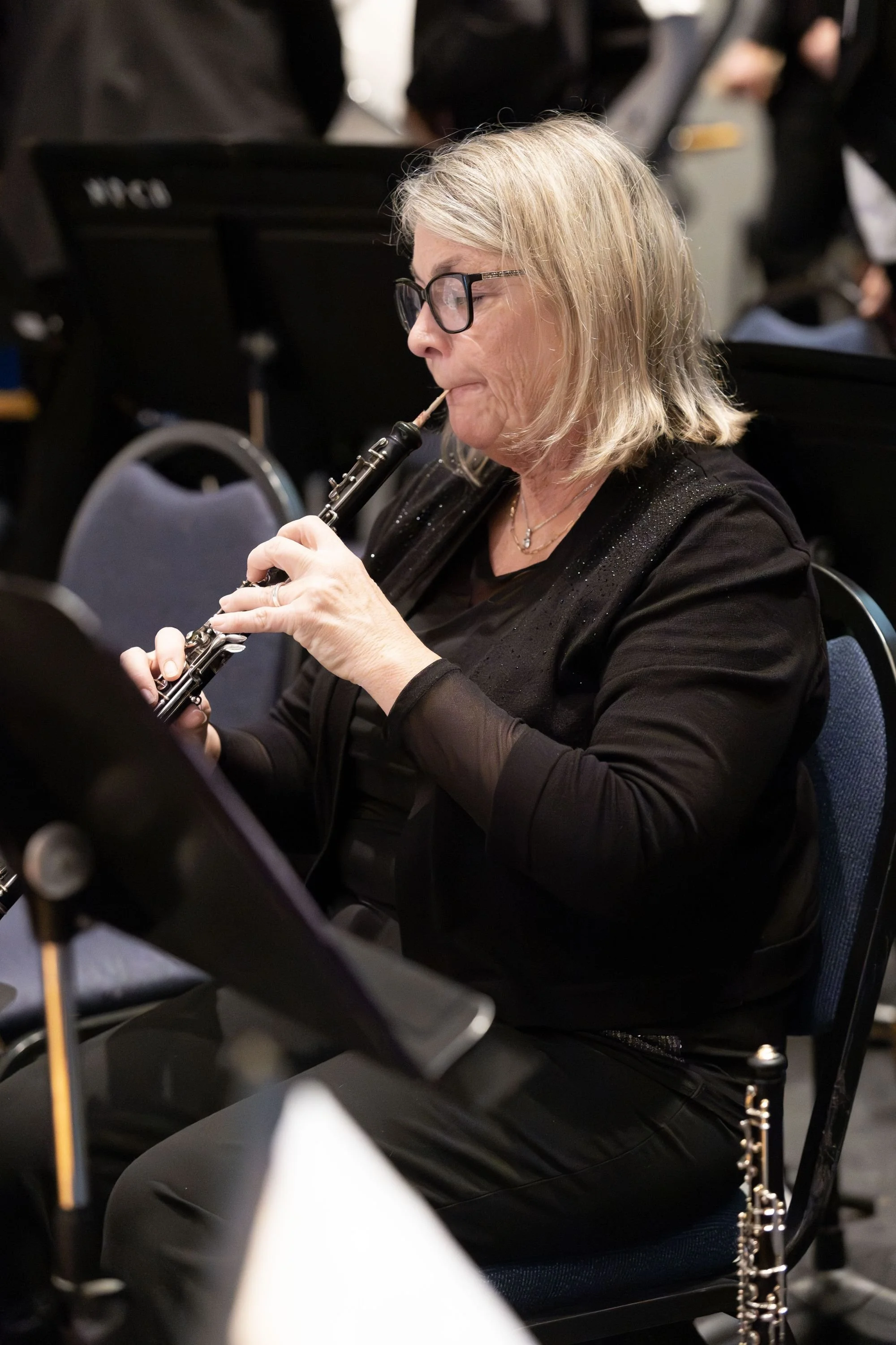 A woman with blond hair, glasses, and black clothing playing a clarinet at an indoor concert.