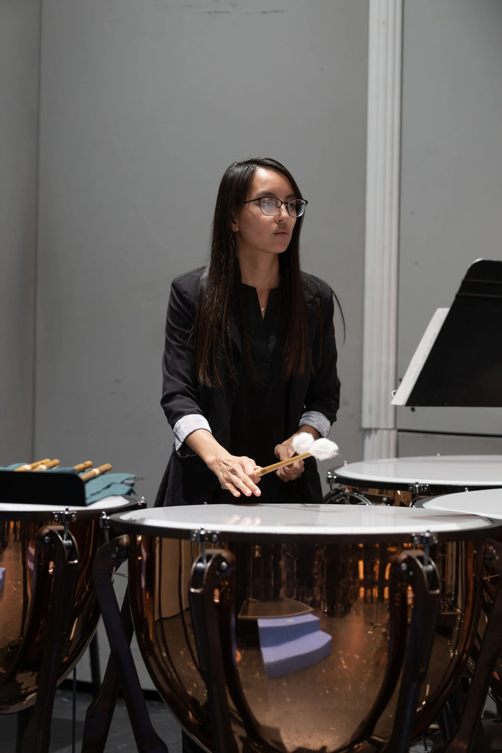 A woman with long dark hair, glasses, and a black jacket playing timpani drums with mallets, standing in a music rehearsal space.