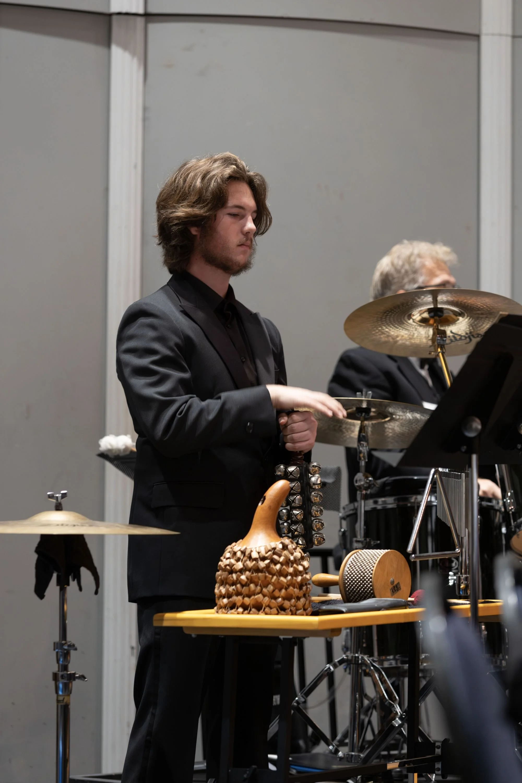 Young male musician in black suit playing percussion instruments during a performance, with a table holding a wooden gourd-shaped shaker and a percussion instrument, behind an older drummer with a cymbal.