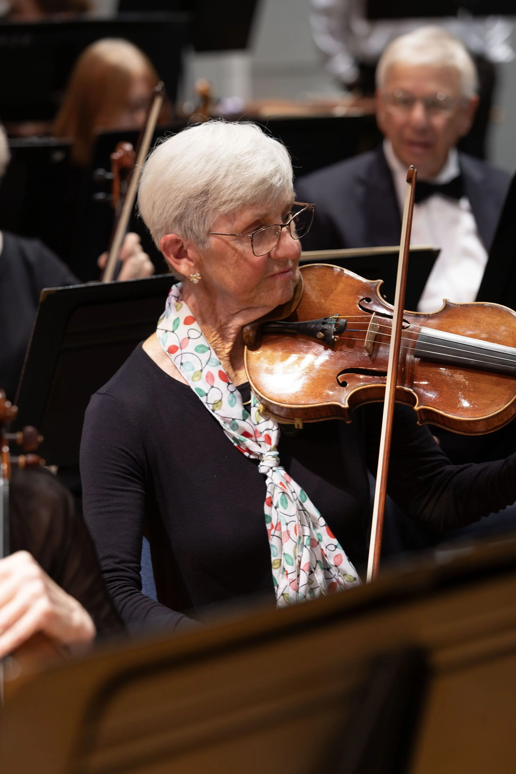 An elderly woman with gray hair, glasses, and a colorful scarf playing the violin in an orchestra.