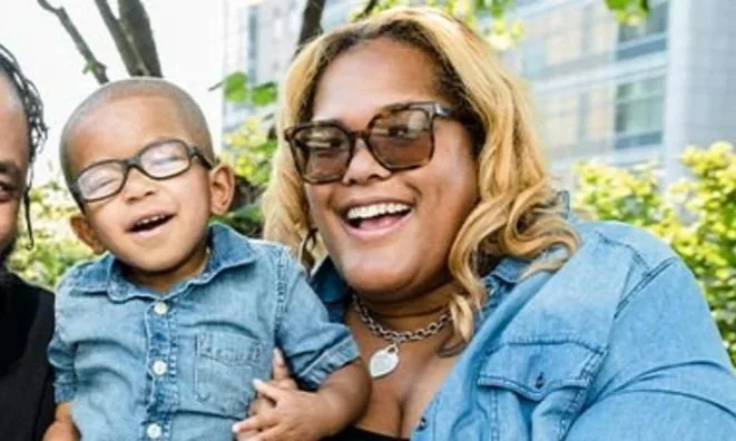 A woman with blonde hair and sunglasses holding a young boy with glasses, both smiling outdoors with trees and buildings in the background.