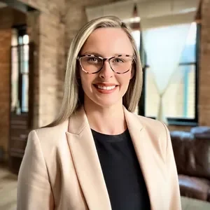 A smiling woman with blonde hair and glasses, wearing a beige blazer and black top, in a well-lit indoor setting with large windows.