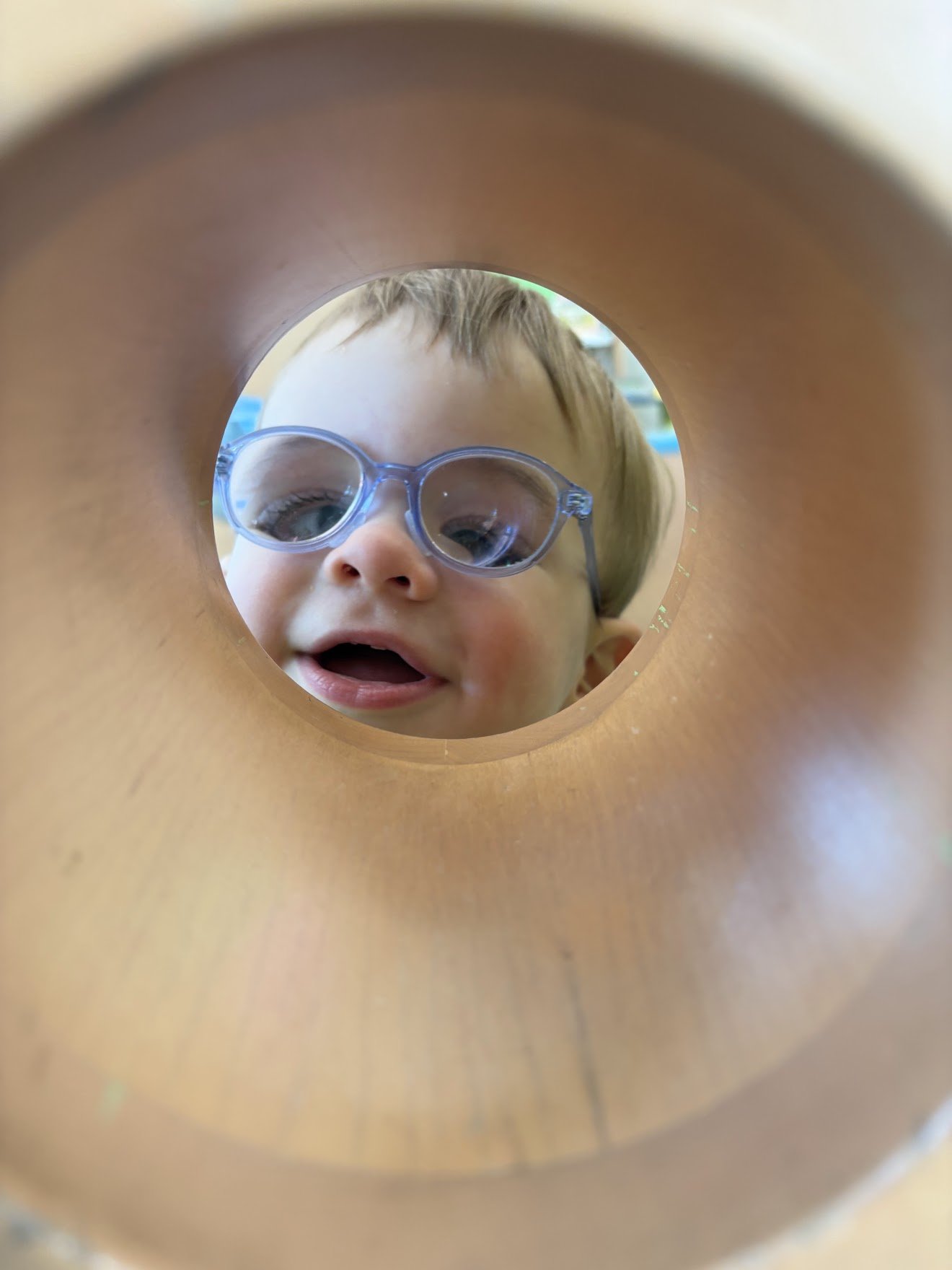 A young boy with glasses looking through a circular hole, with his face partially visible and his mouth open.