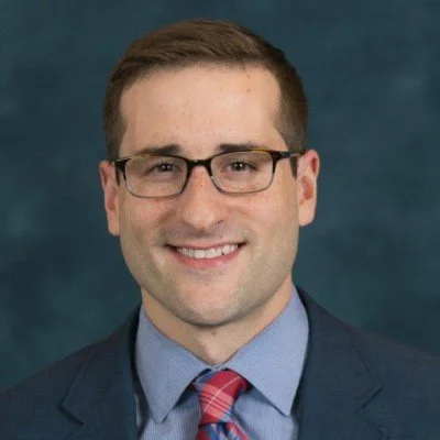 A man with short brown hair and glasses, wearing a suit, blue shirt, and pink tie, smiling against a dark background.