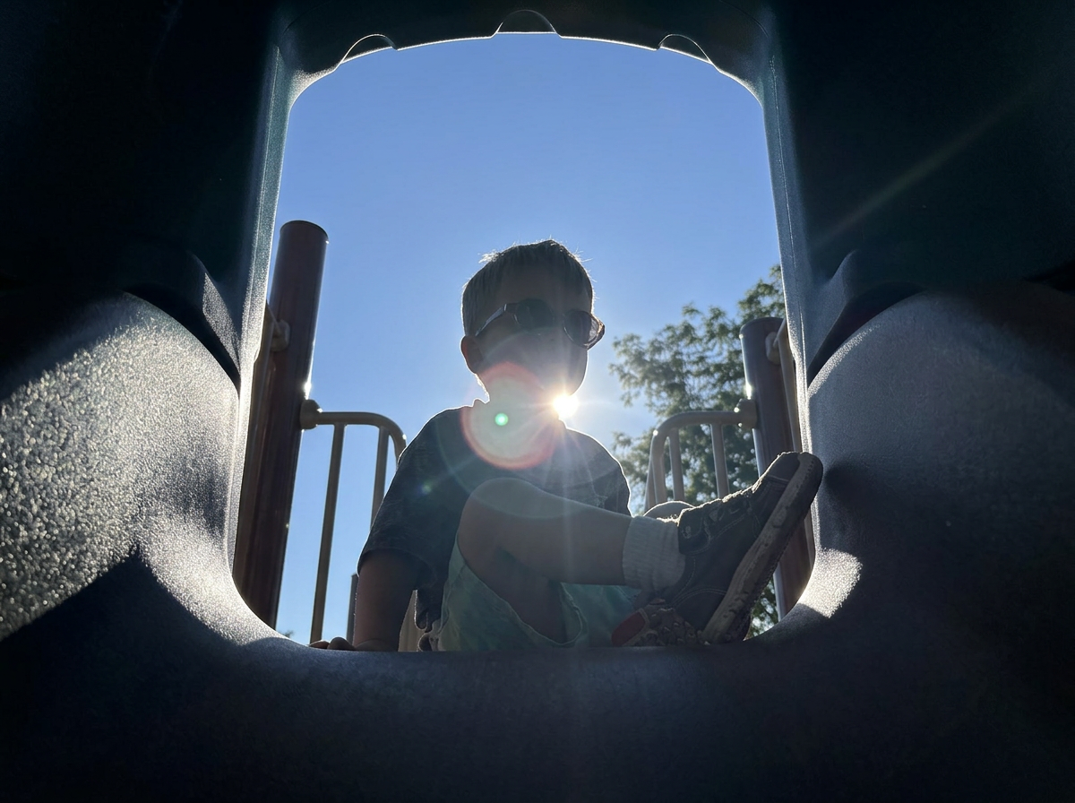 Child wearing sunglasses sitting on playground steps with the sky and trees in the background, seen from inside a play structure.