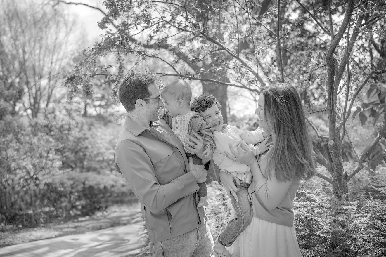 A family of four smiling and enjoying time together outdoors in a park, with trees and foliage in the background. The father is holding a toddler, and the mother is holding a young boy.