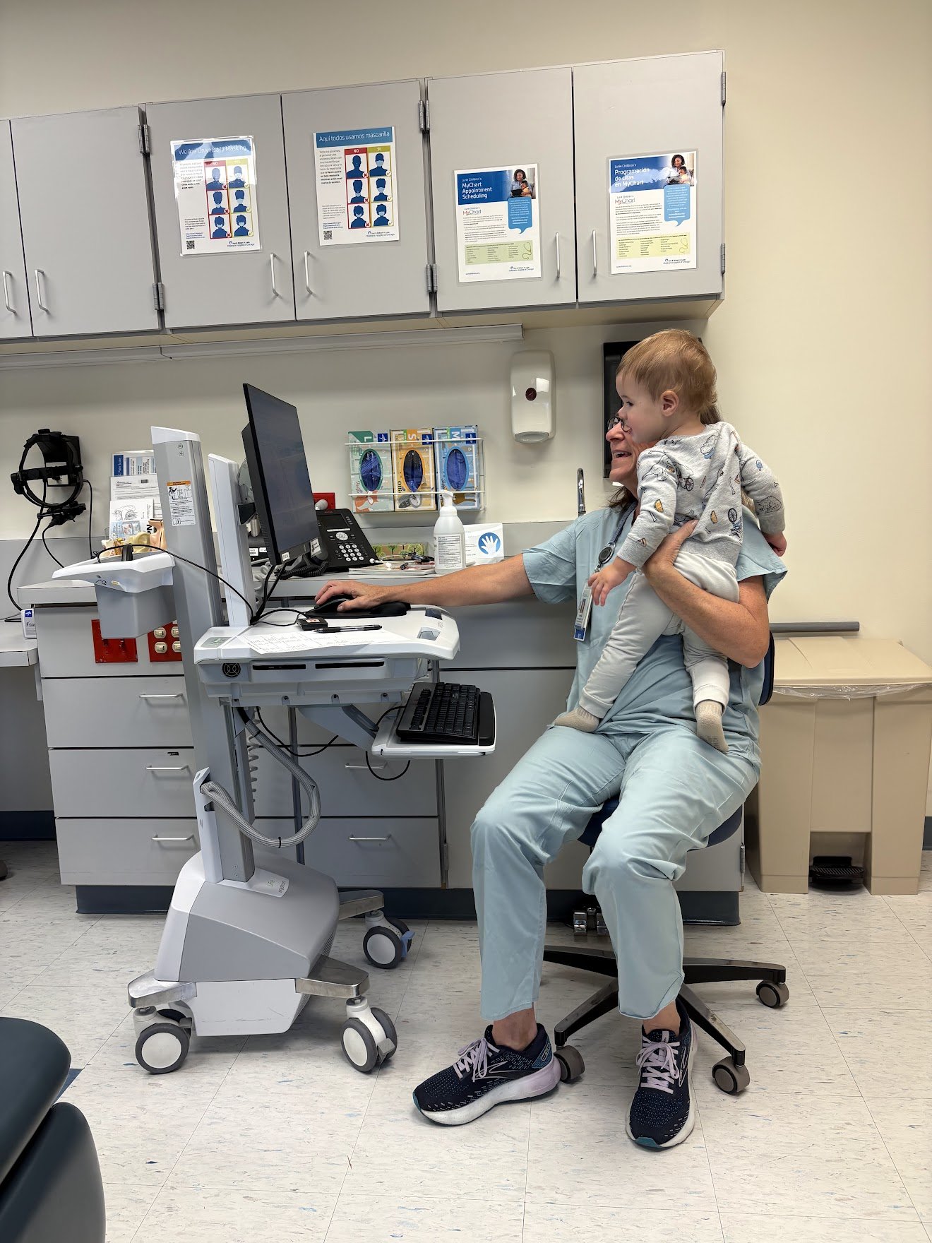 A woman doctor sitting on a rolling stool in a medical office, holding a small child on her lap, while working on a computer. The woman is smiling, and the child is looking to the side. Medical equipment and cabinets are visible in the background.