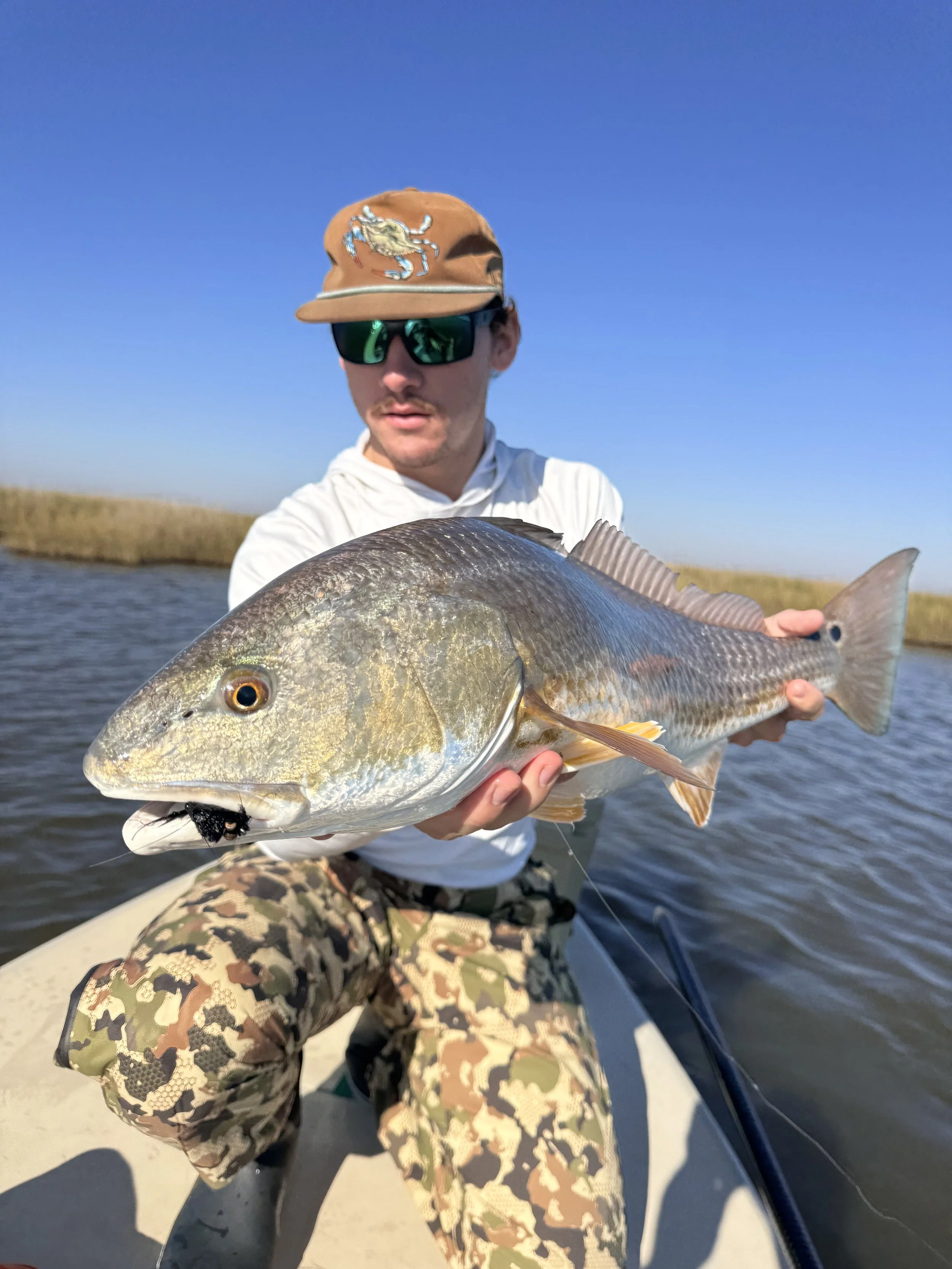 Man wearing camouflage pants, a white long-sleeve shirt, sunglasses, and a tan hat with an embroidered crab, holding a large fish on a boat in a waterway with grassy banks under a clear blue sky.