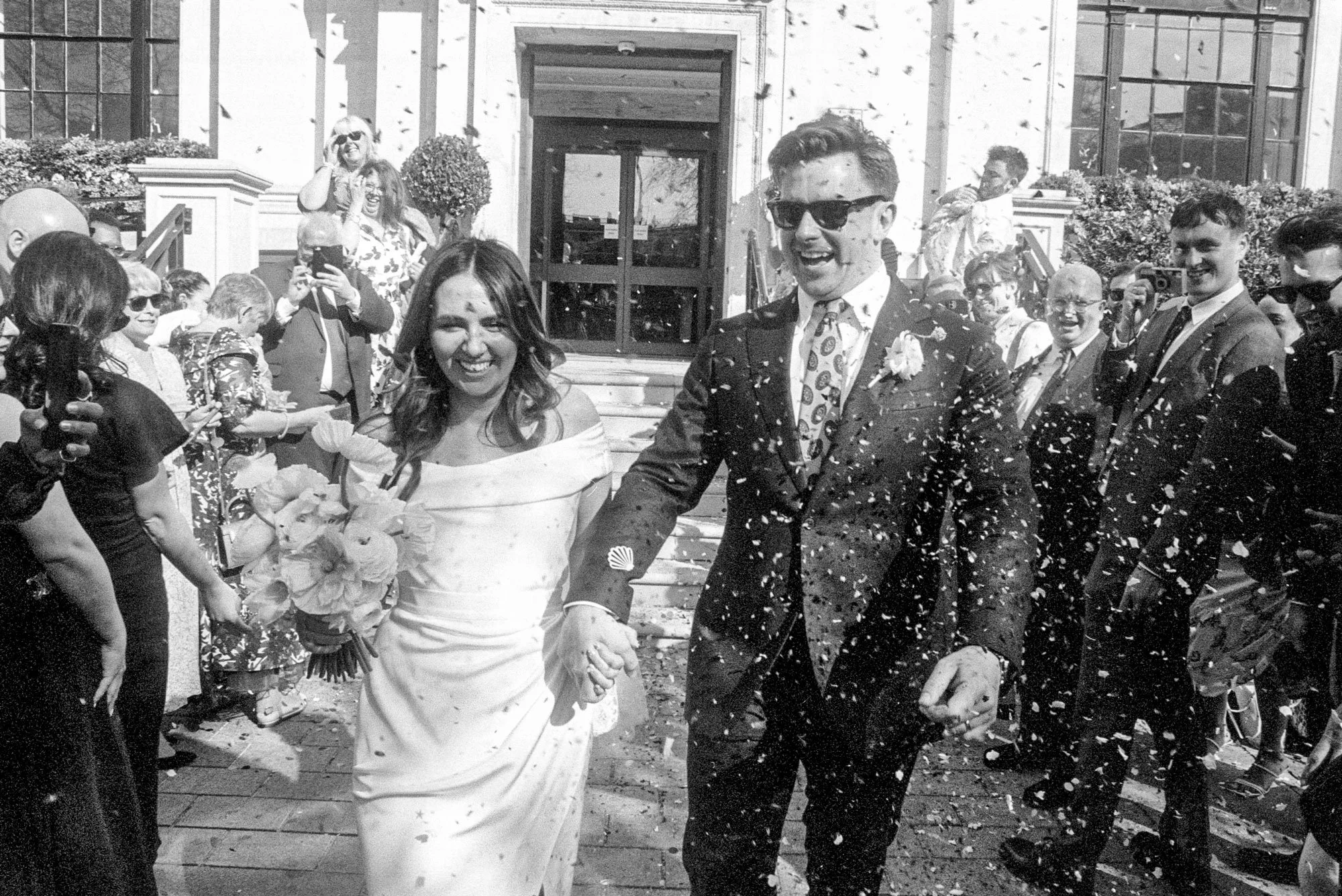 A black-and-white photo of a wedding celebration taking place outdoors, featuring a smiling bride and groom holding hands, surrounded by guests throwing confetti.