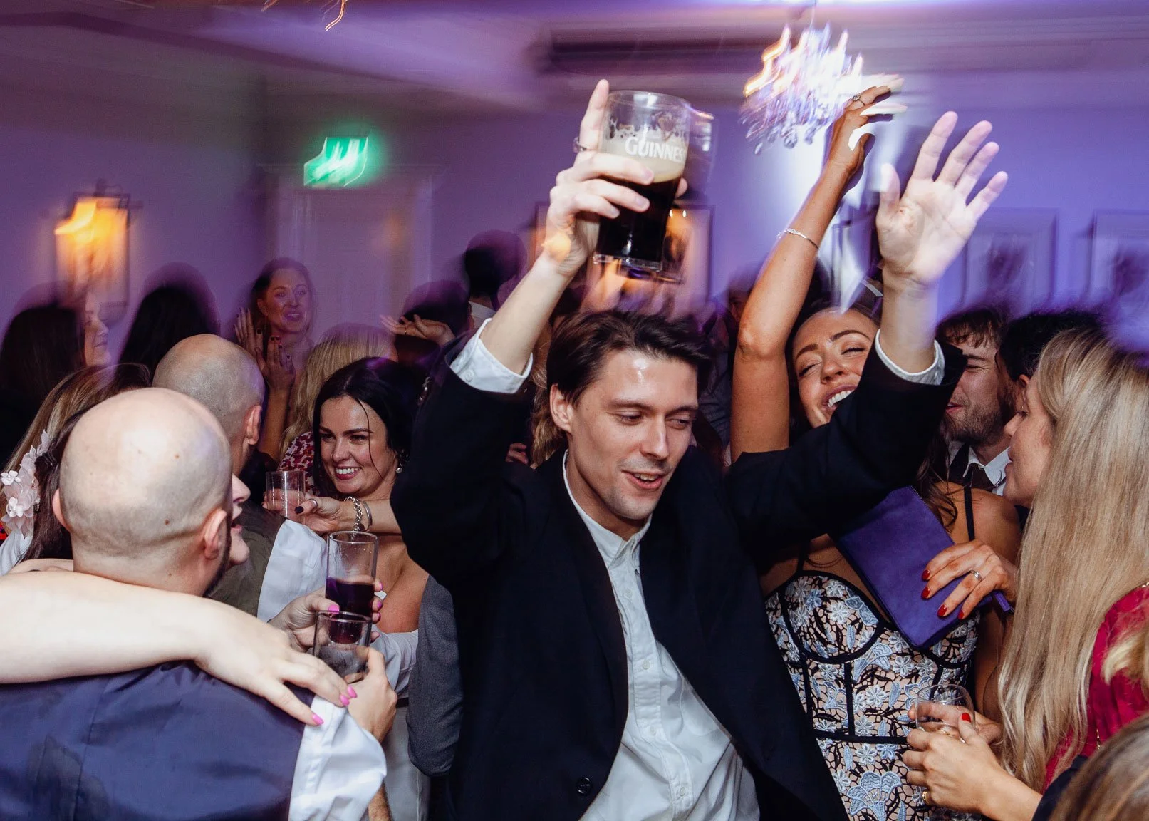 People celebrating and dancing at a party, with one man holding a pint of Guinness beer and smiling, surrounded by others enjoying drinks and festive atmosphere.