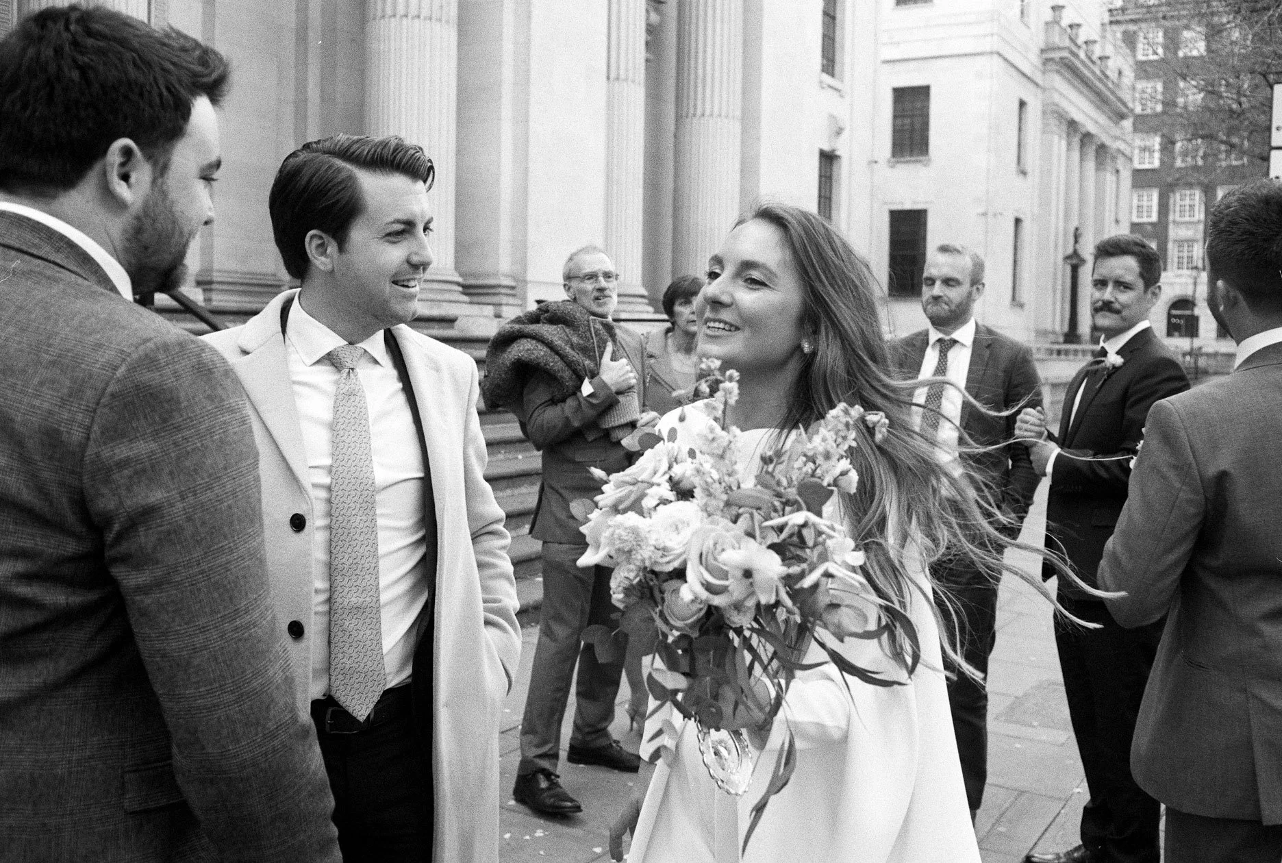 A woman in a white dress holding a bouquet of flowers is smiling and interacting with men dressed in formal suits outside a historic building with steps.