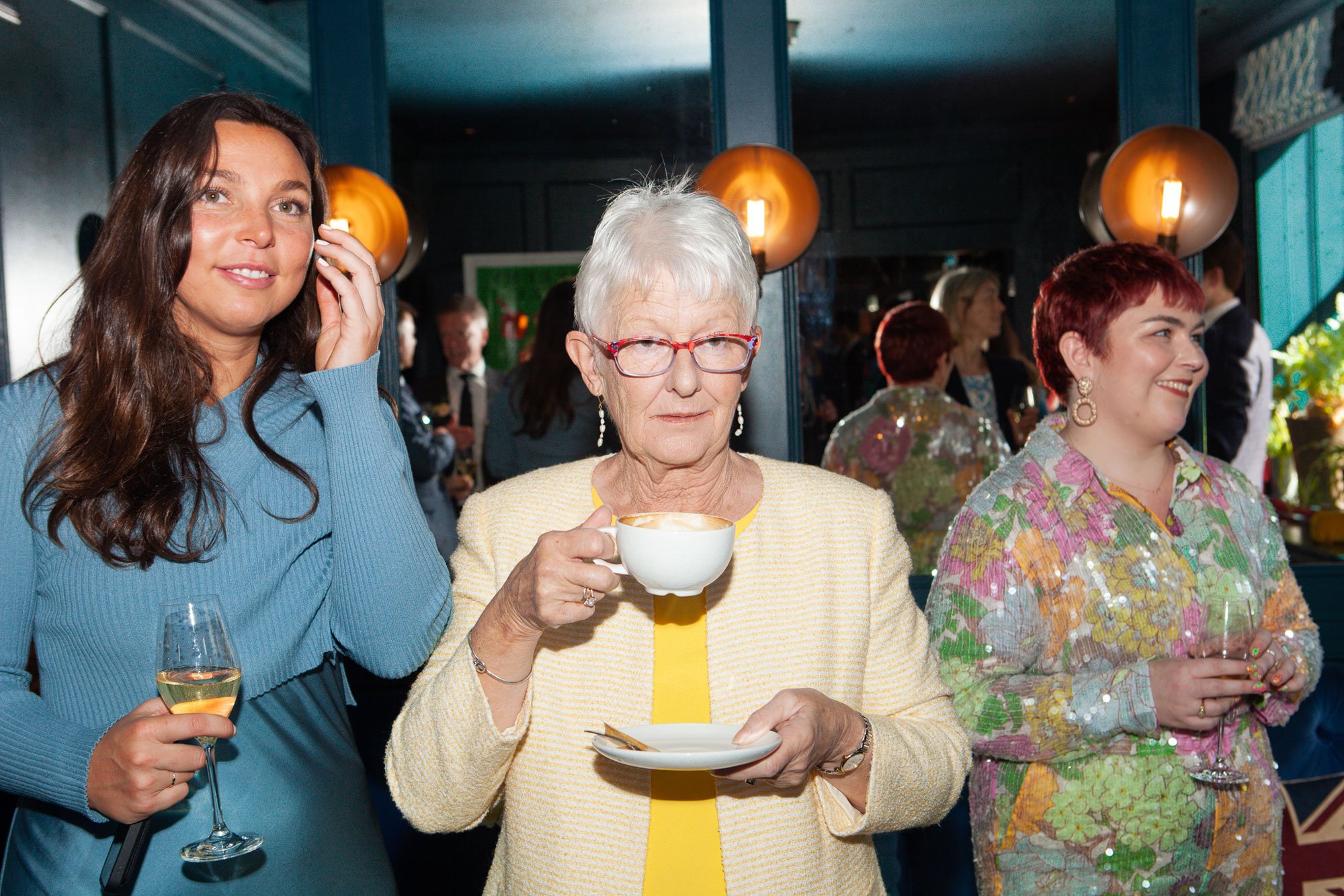 Three women at a social gathering, with the woman in the middle holding a cup of coffee or tea, the woman on the left holding a glass of white wine, and the woman on the right holding a glass of champagne, in a dimly lit indoor setting.