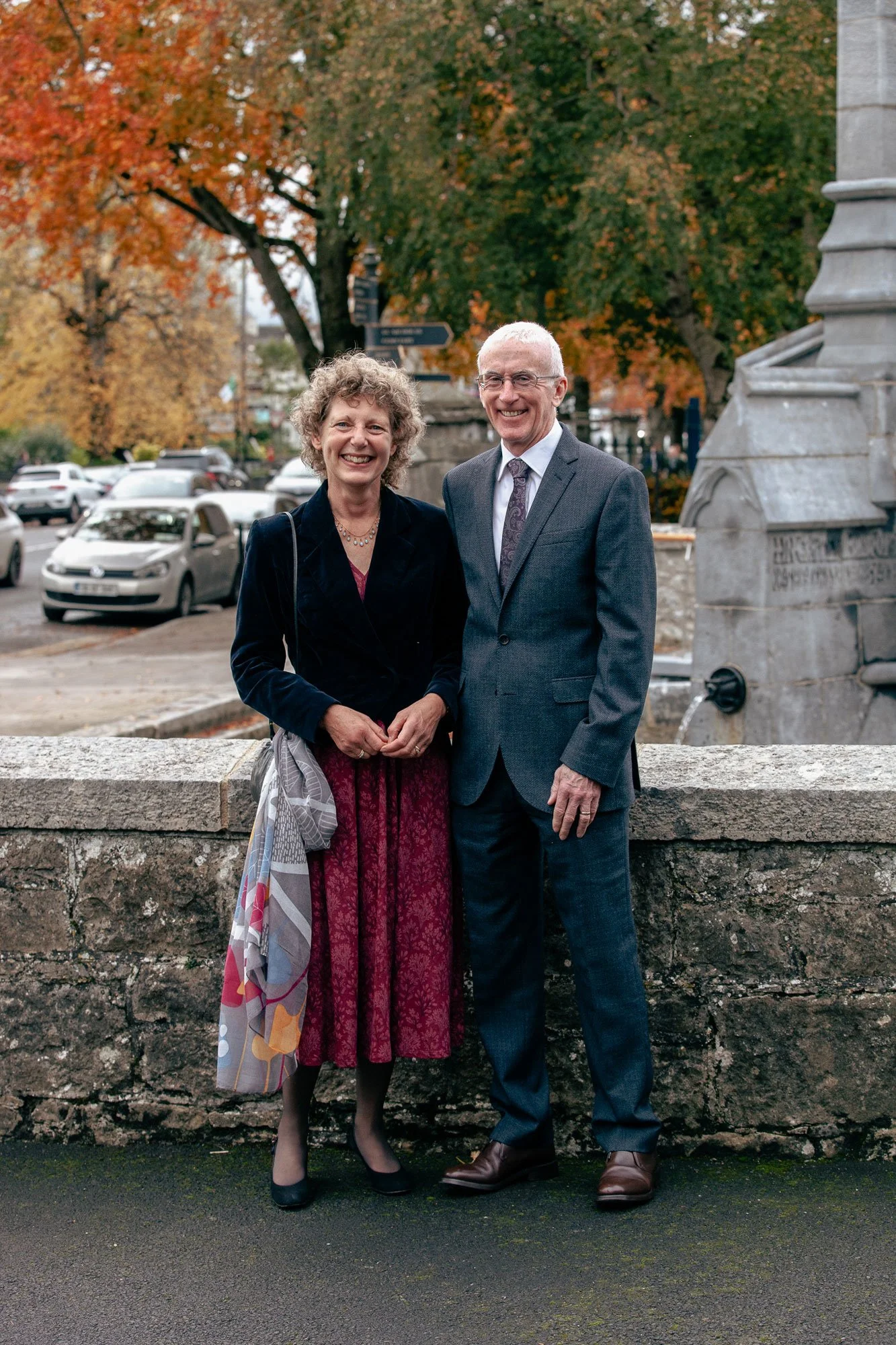 A man and woman dressed formally, standing outdoors on a stone ledge, smiling. The woman has curly hair, dark blazer, patterned red skirt, black shoes, and a patterned bag. The man has short white hair, glasses, a gray suit, patterned tie, and brown shoes. Behind them are autumn trees, parked cars, and part of a stone monument or fountain.