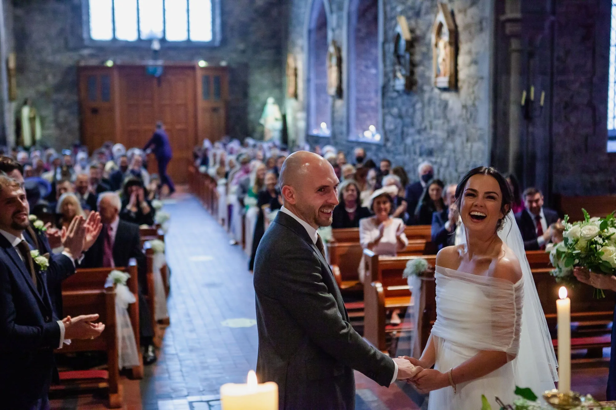Bride and groom holding hands during their wedding ceremony inside a church, with guests applauding in the background.