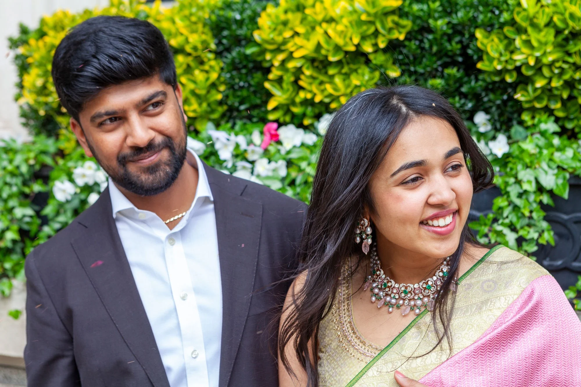 A man and woman smiling, dressed formally, standing outdoors with green bushes and flowers in the background.