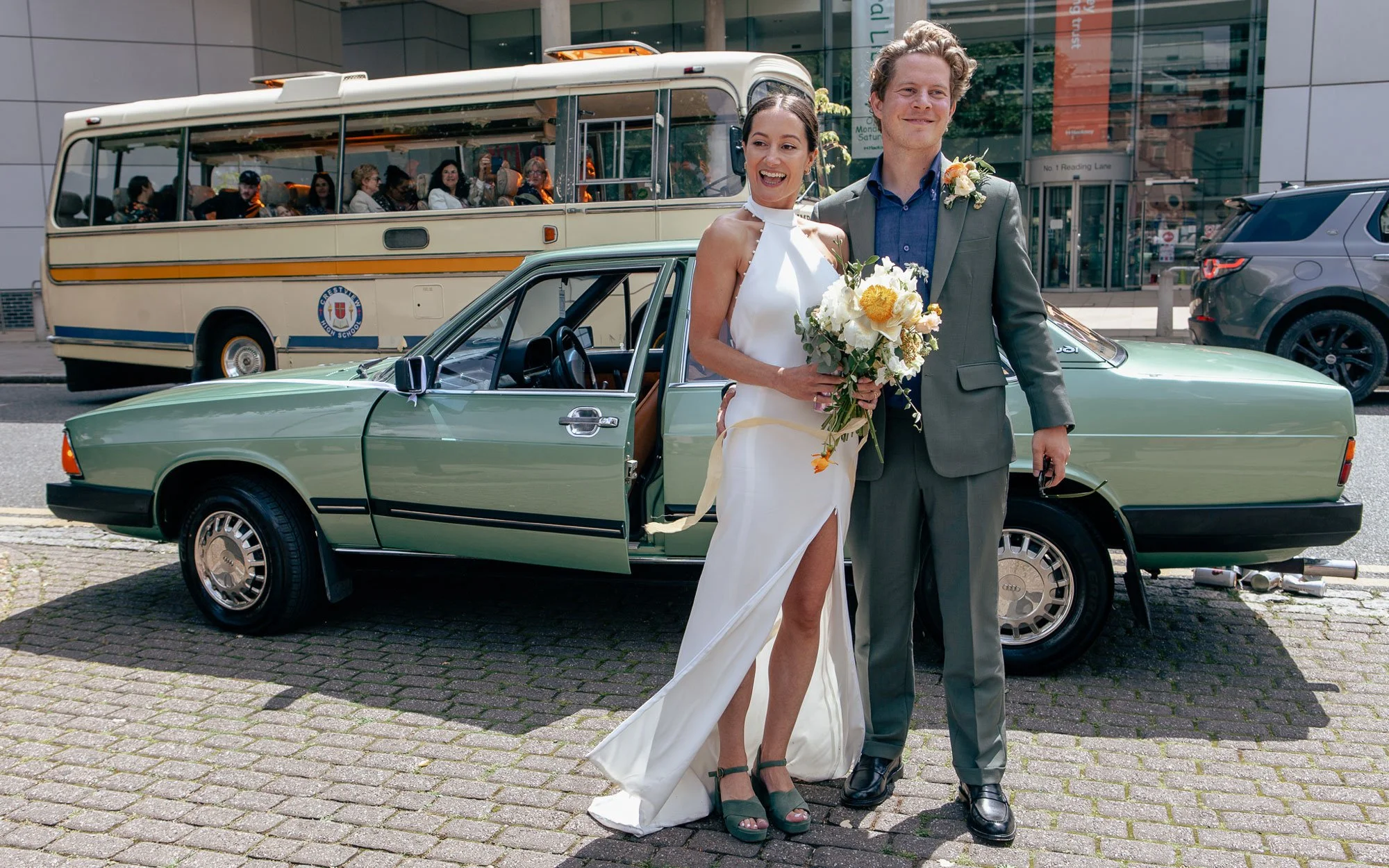 A newly married couple stands in front of a vintage green car, smiling and holding flowers and a car key, with a bus and city buildings in the background.