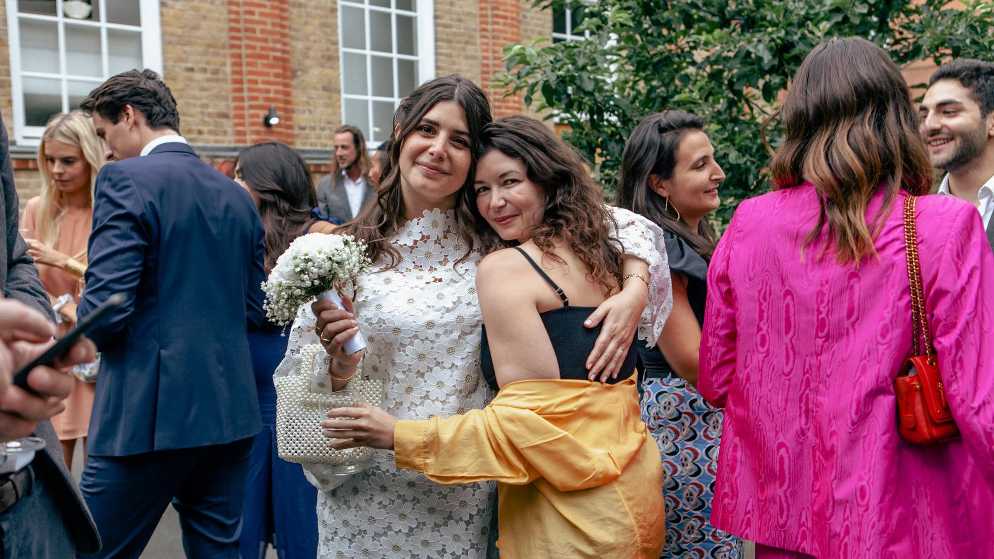 Two women with brown curly hair, one holding a white flower bouquet, hugging at an outdoor event with other people in colorful suits and dresses in the background.