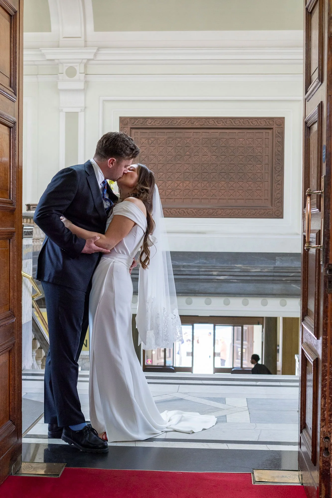 A newlywed couple sharing a kiss inside a grand building with high ceilings, ornate architecture, and large windows in the background.