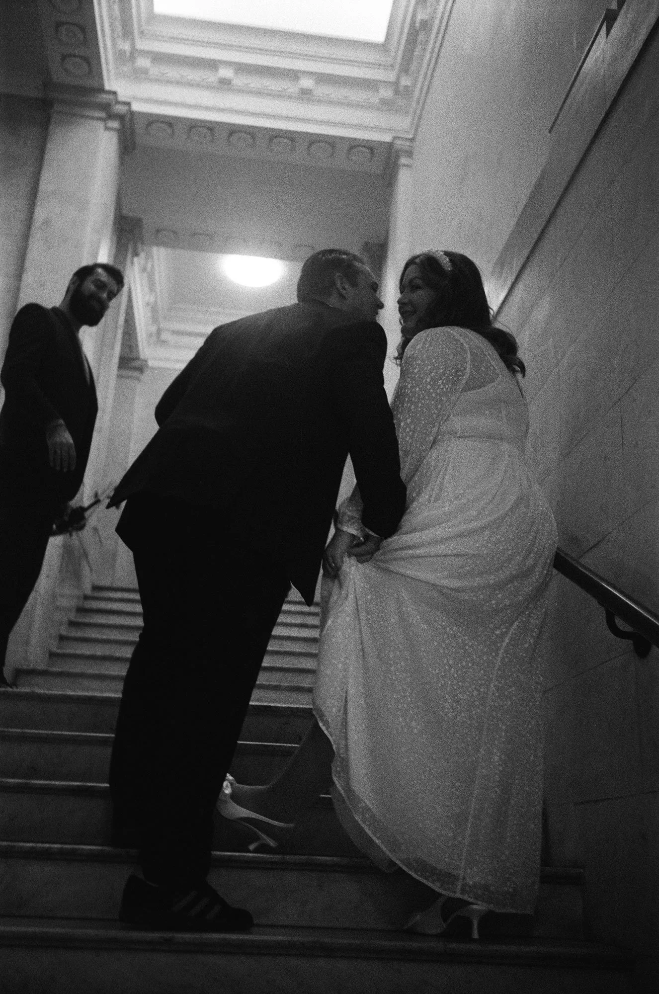 A black and white photo of a couple standing on stairs, smiling and leaning towards each other, with a woman holding up her gown. Another man stands to the side, holding something in his hand, in a grand indoor setting with ornate ceiling details.