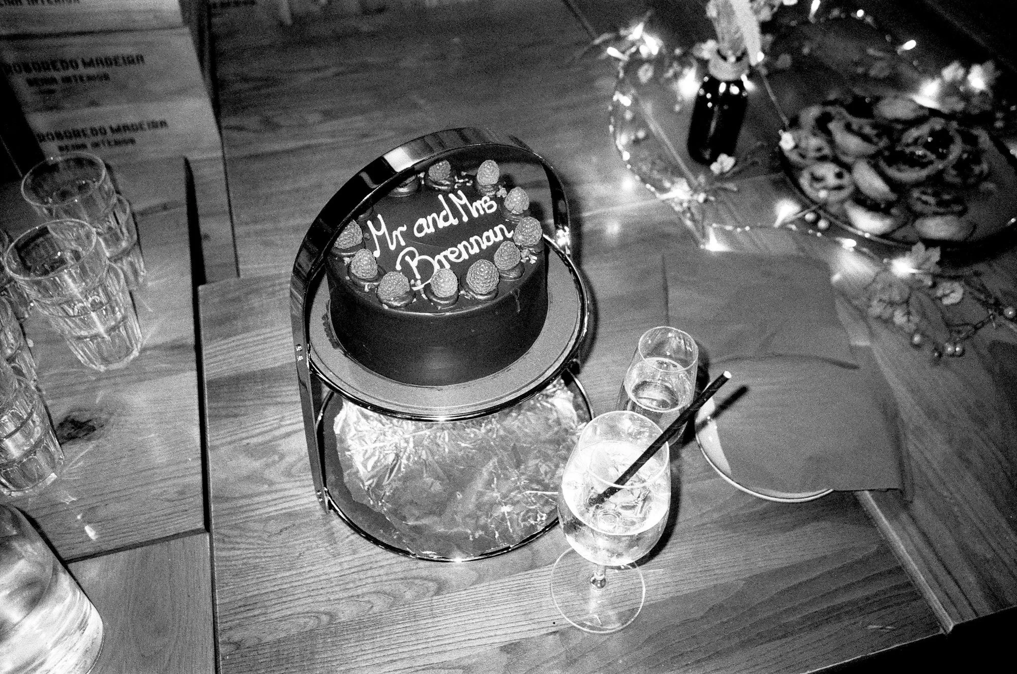 A cake with the message "Mr and Mrs Brennan" on top, surrounded by strawberries, on a table with glasses, cookies, and decorations, at a celebration or wedding reception.