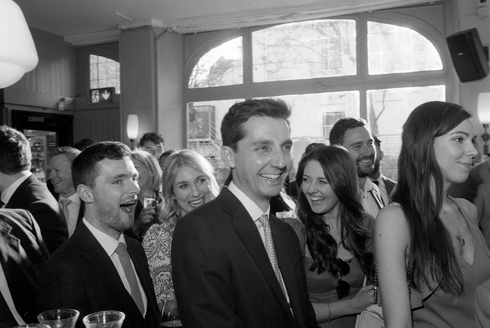 Black and white photo of a group of smiling people at a social gathering or party, with large windows in the background letting in natural light.