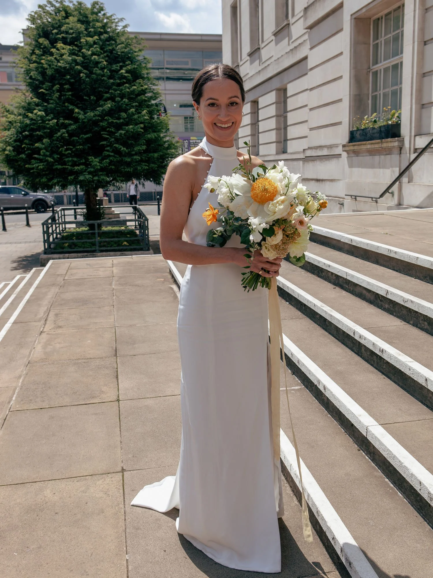 A woman in a white dress holding a bouquet of flowers, standing outside on steps in front of a building and trees, smiling.