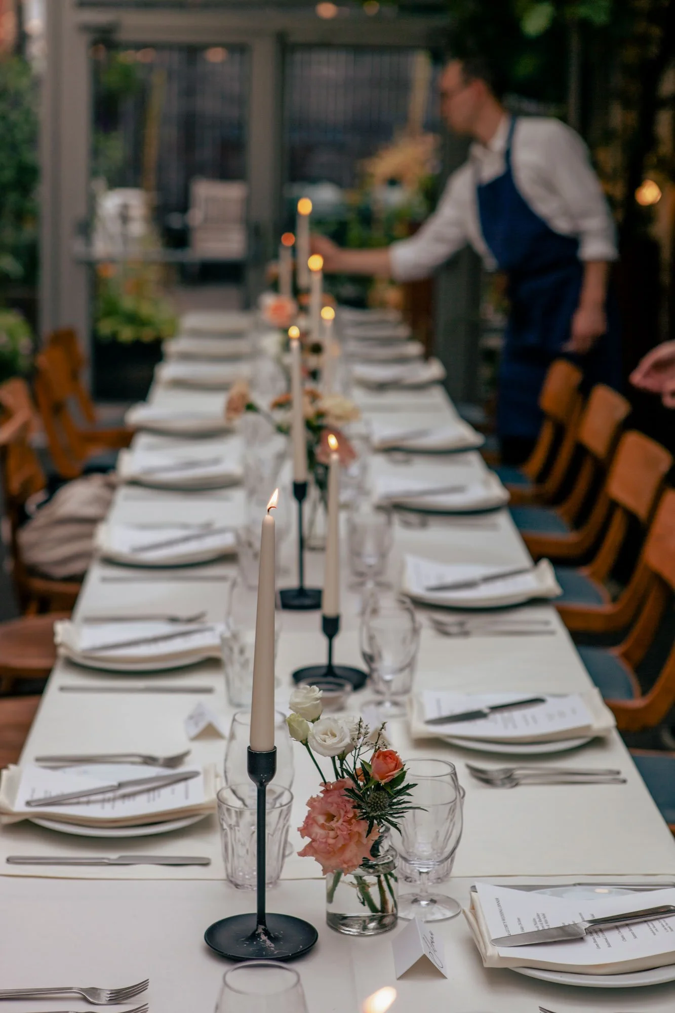 A long dining table set for a formal event with candles, flowers, and glassware. A person in a white shirt and blue apron is in the background, preparing or serving. The scene appears to be outdoors or in a conservatory with greenery.