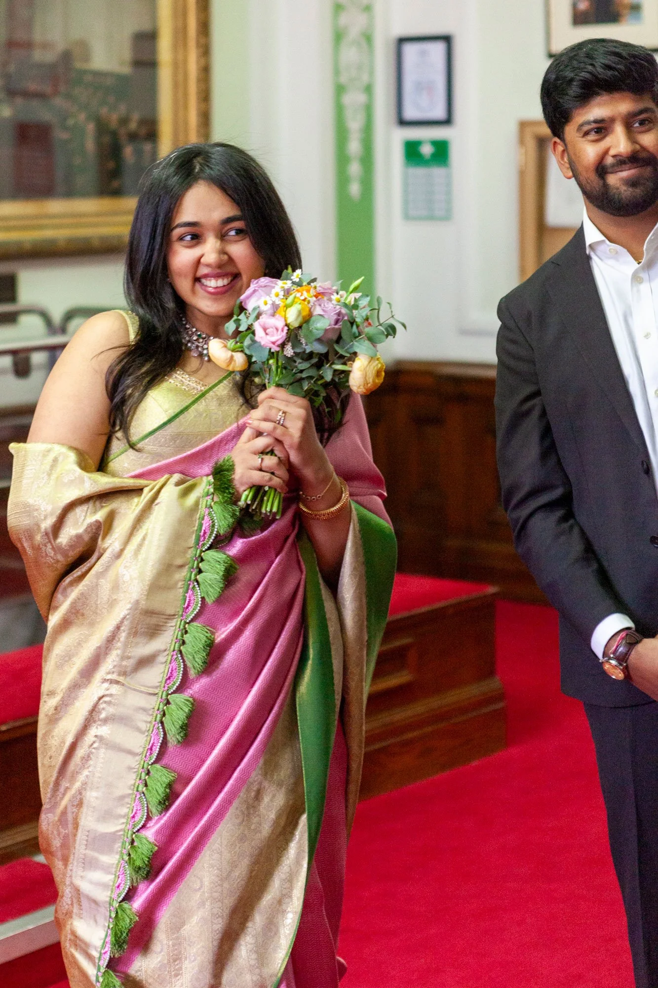 An Indian woman wearing a gold, pink, and green saree holding a bouquet of flowers, smiling in a formal indoor setting with a red carpet and wooden paneling.