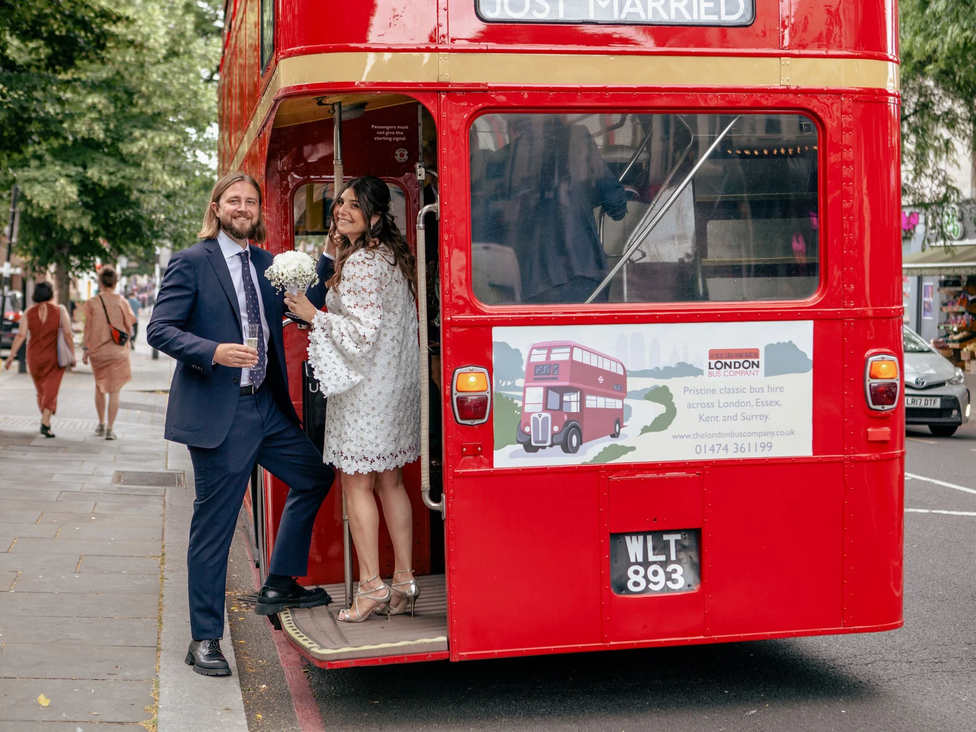 A groom and bride in wedding attire standing on the steps of a red double-decker bus, smiling and holding a bouquet of white flowers, in an urban street setting with pedestrians and parked cars.