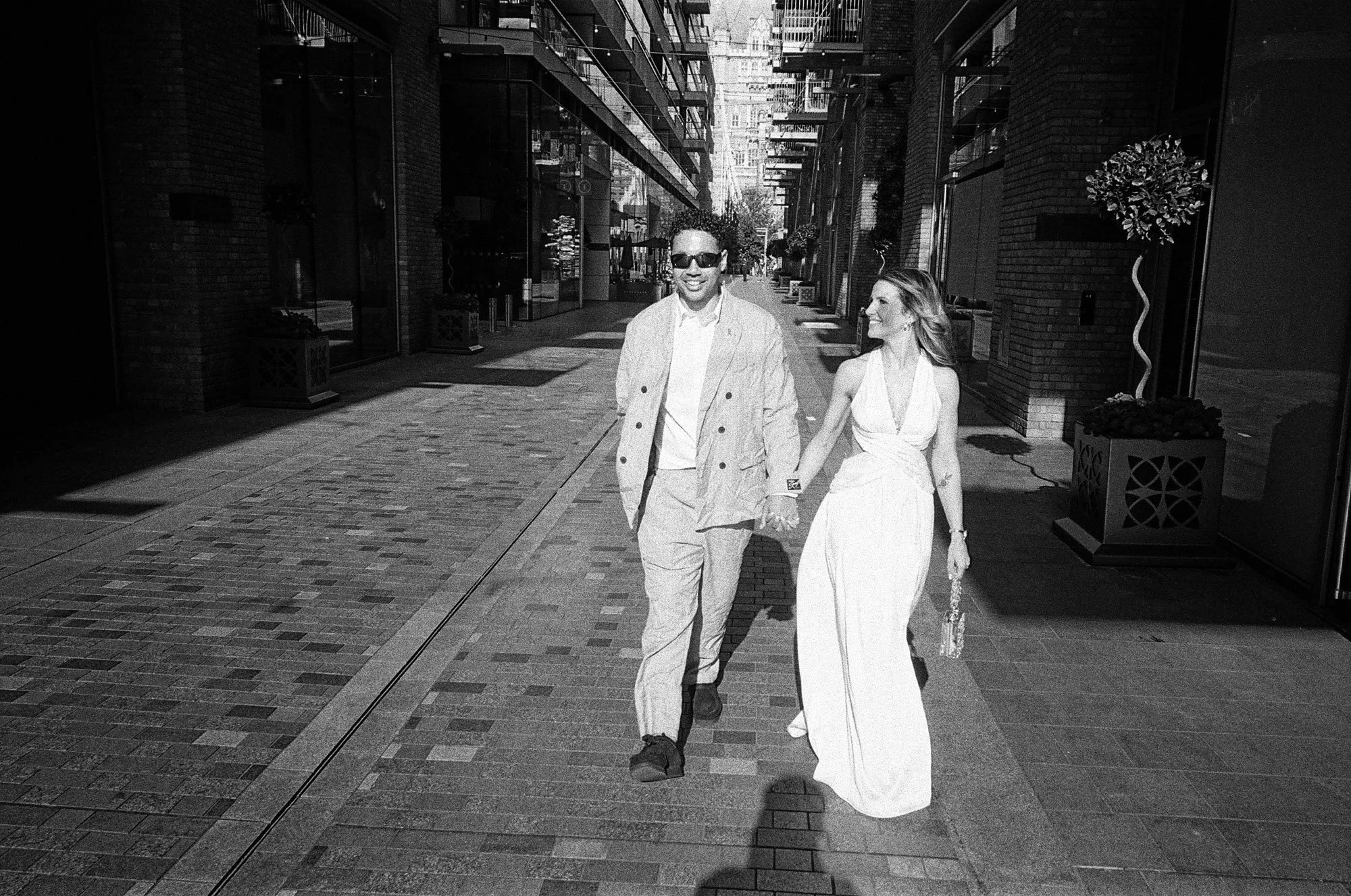 A man and woman holding hands and walking on a city street. The man is wearing sunglasses, a light-colored suit, and the woman is wearing a long, sleeveless dress. They are smiling and looking at each other.