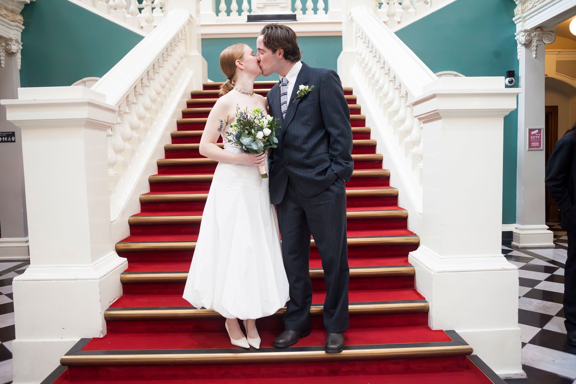 A bride and groom sharing a kiss on a red-carpeted staircase inside a building with teal walls and white ornate trim.