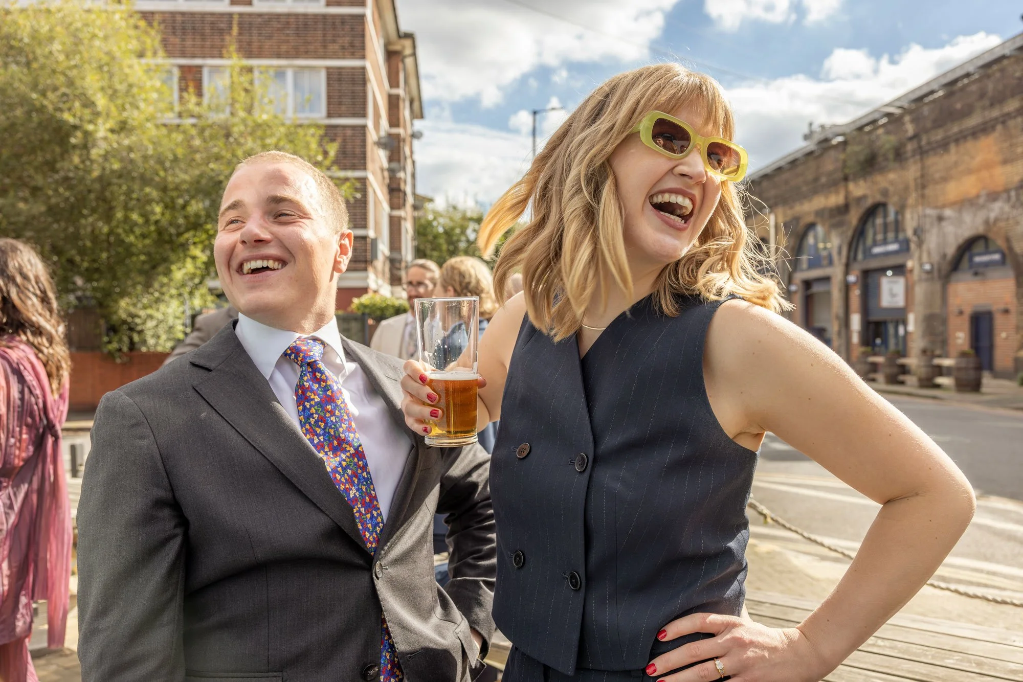A man in a gray suit and a woman in a black sleeveless top laughing outdoors, with the woman holding a beer glass. The background shows a street scene with brick buildings, trees, and a partly cloudy sky.
