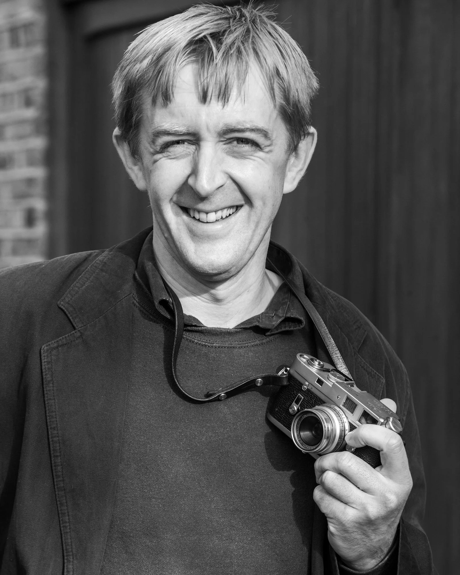A smiling man holding a vintage camera, wearing a dark jacket and shirt, with a strap around his neck, in black and white.