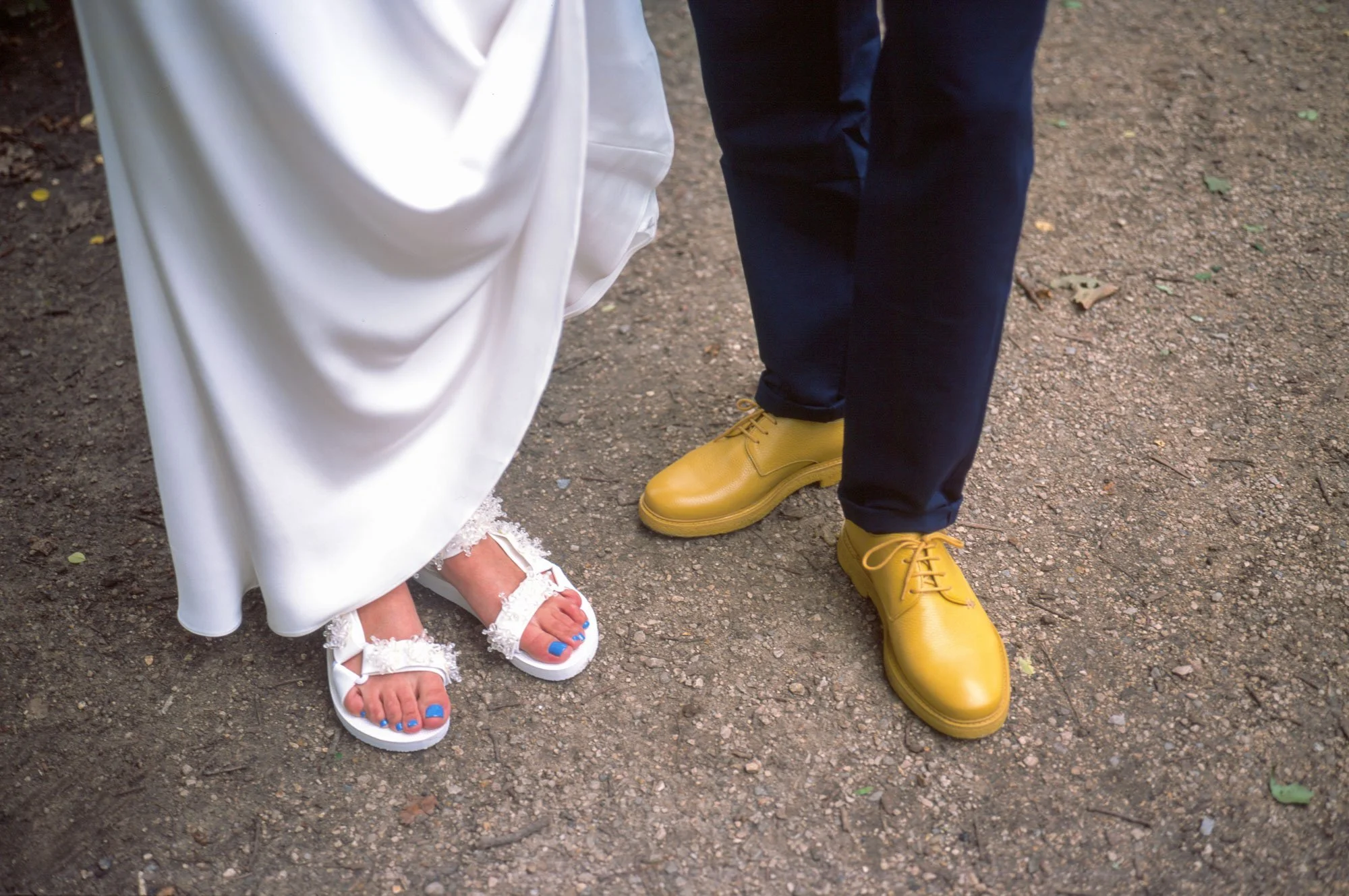 Close-up of two people standing on a dirt path, one wearing a white dress and white sandals with blue toes visible, the other wearing navy pants and bright yellow shoes.