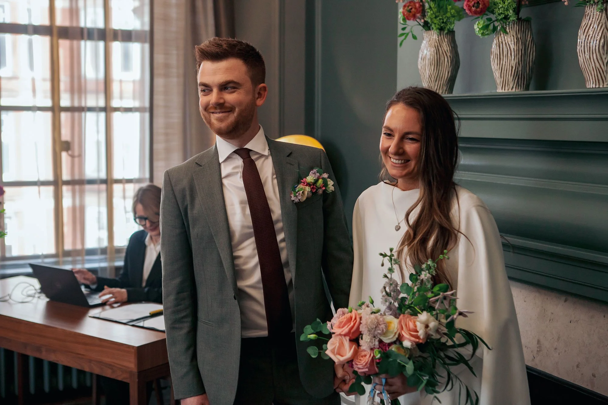 A smiling man in a gray suit with a boutonniere and a woman in a white dress holding a bouquet of pink and white flowers, standing indoors with a dark green wall and decorative vases, and a woman working on a laptop in the background.